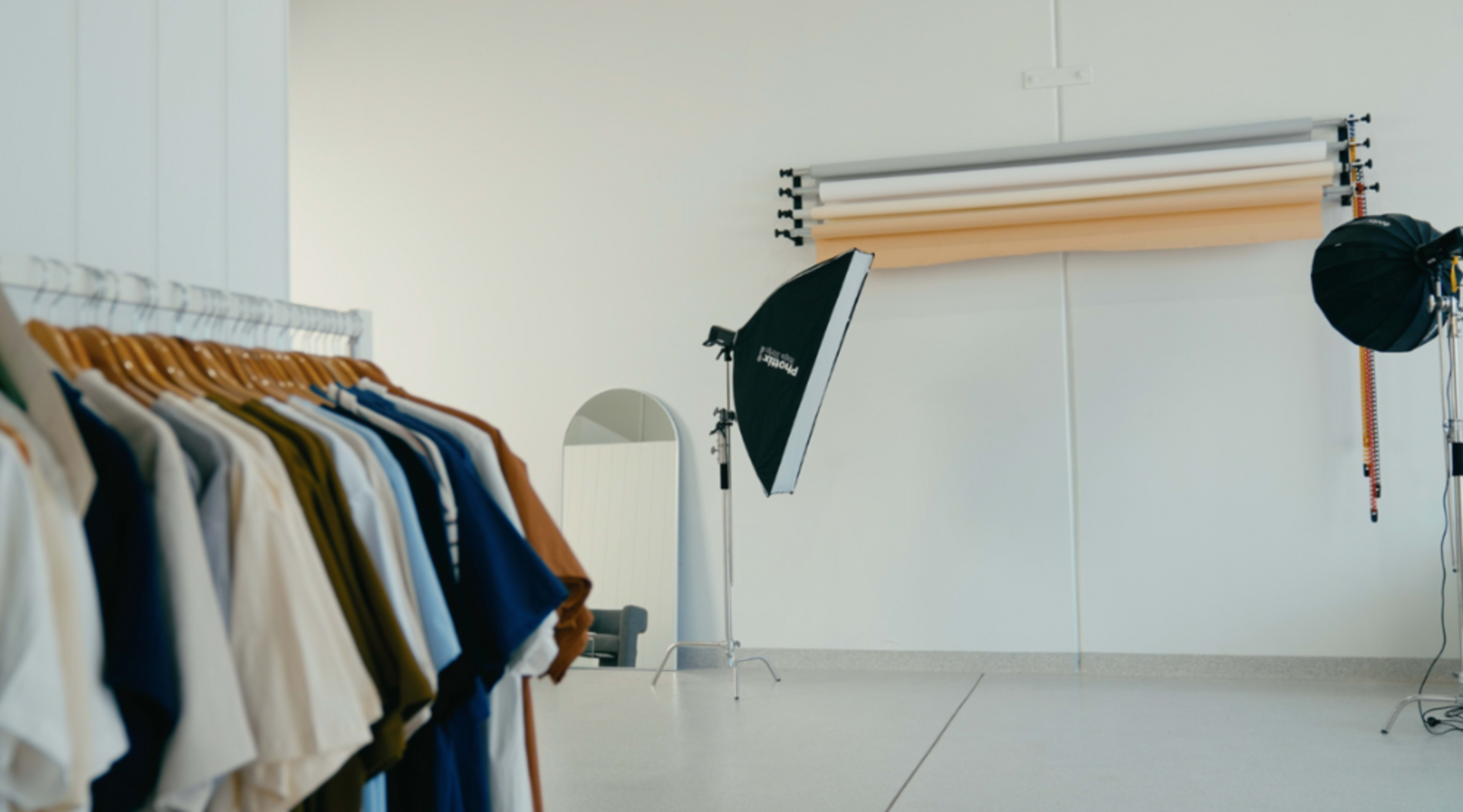 A clothing rack displays various colored shirts next to a photography setup featuring a backdrop and studio lights in a minimalist room.
