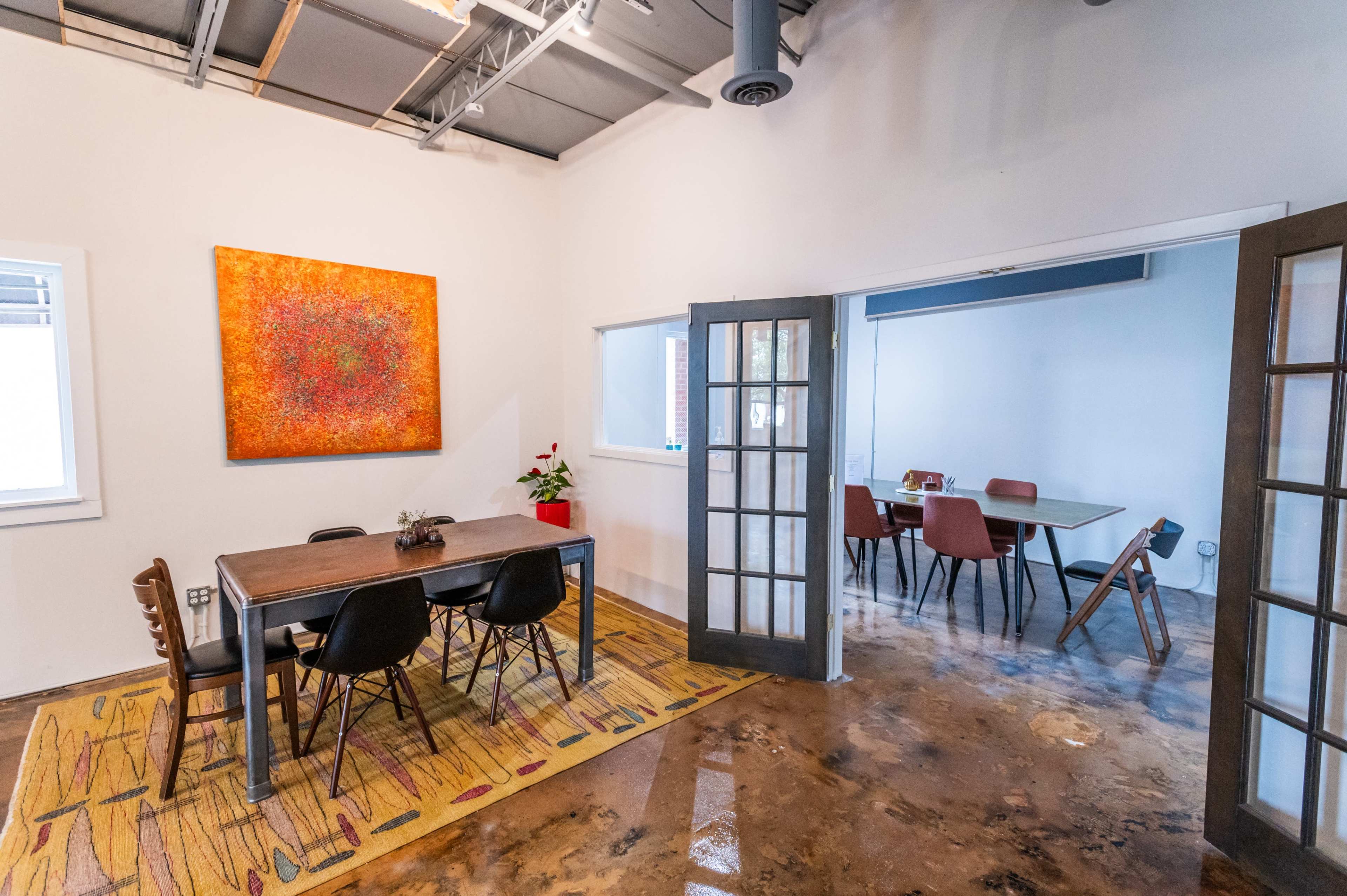 A modern meeting space with a large wooden table surrounded by black chairs, an orange artwork on the wall, and a second room visible through glass doors featuring additional seating.