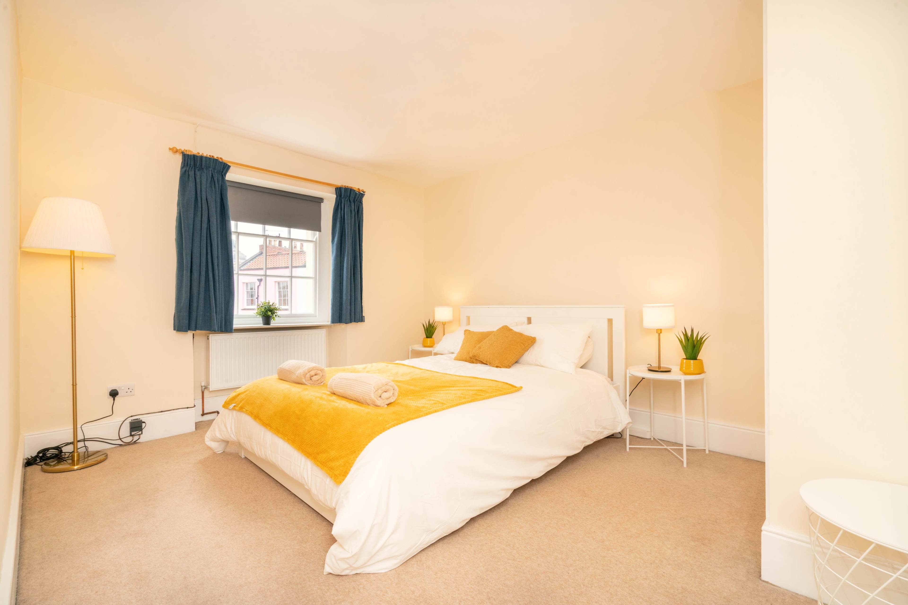A neatly arranged bedroom featuring a large bed with a yellow blanket, two lamps, and a window with blue curtains.