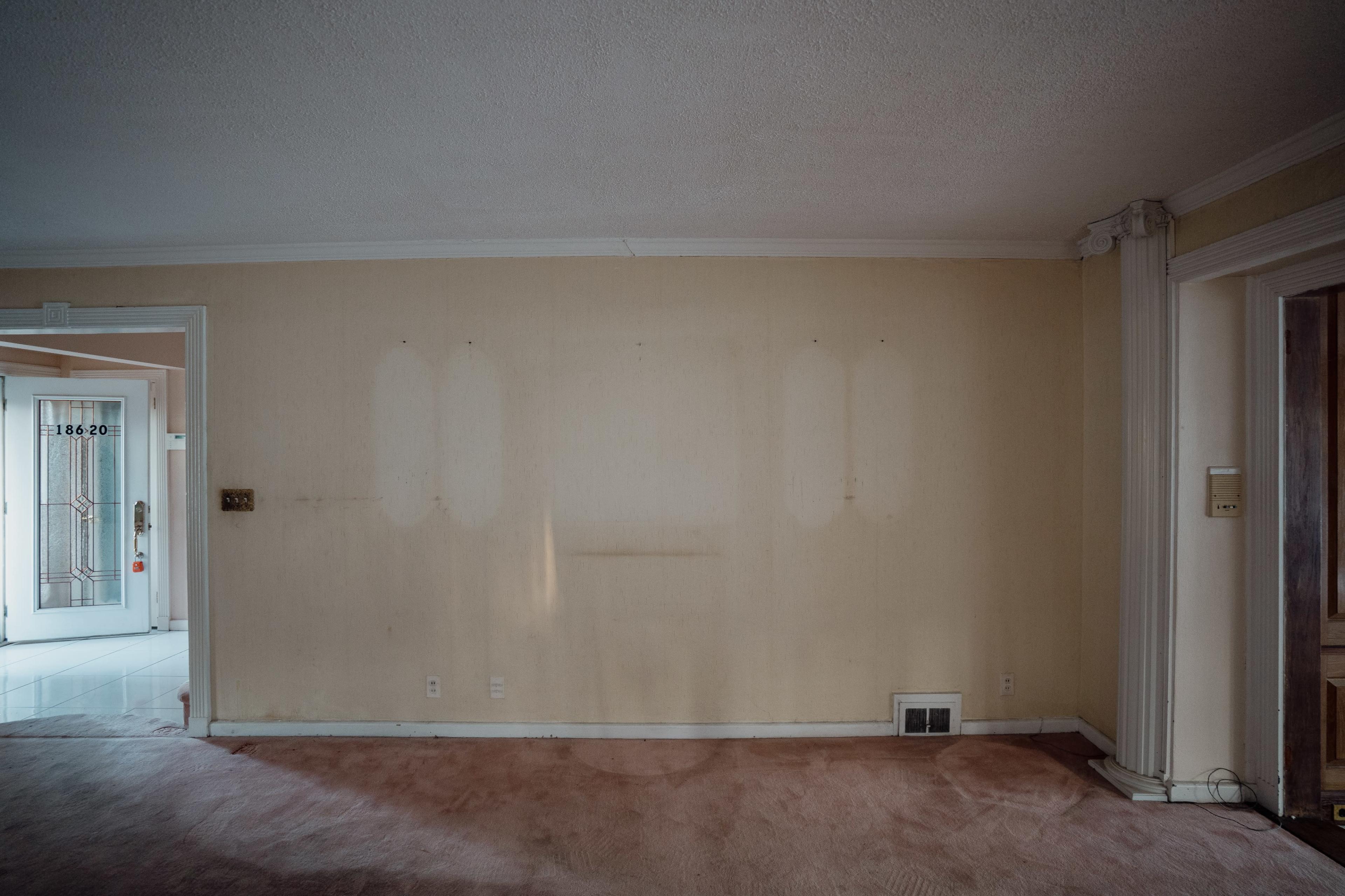 The image shows a vacant living room with beige walls and a carpeted floor, featuring a large window and an entrance door in the background.