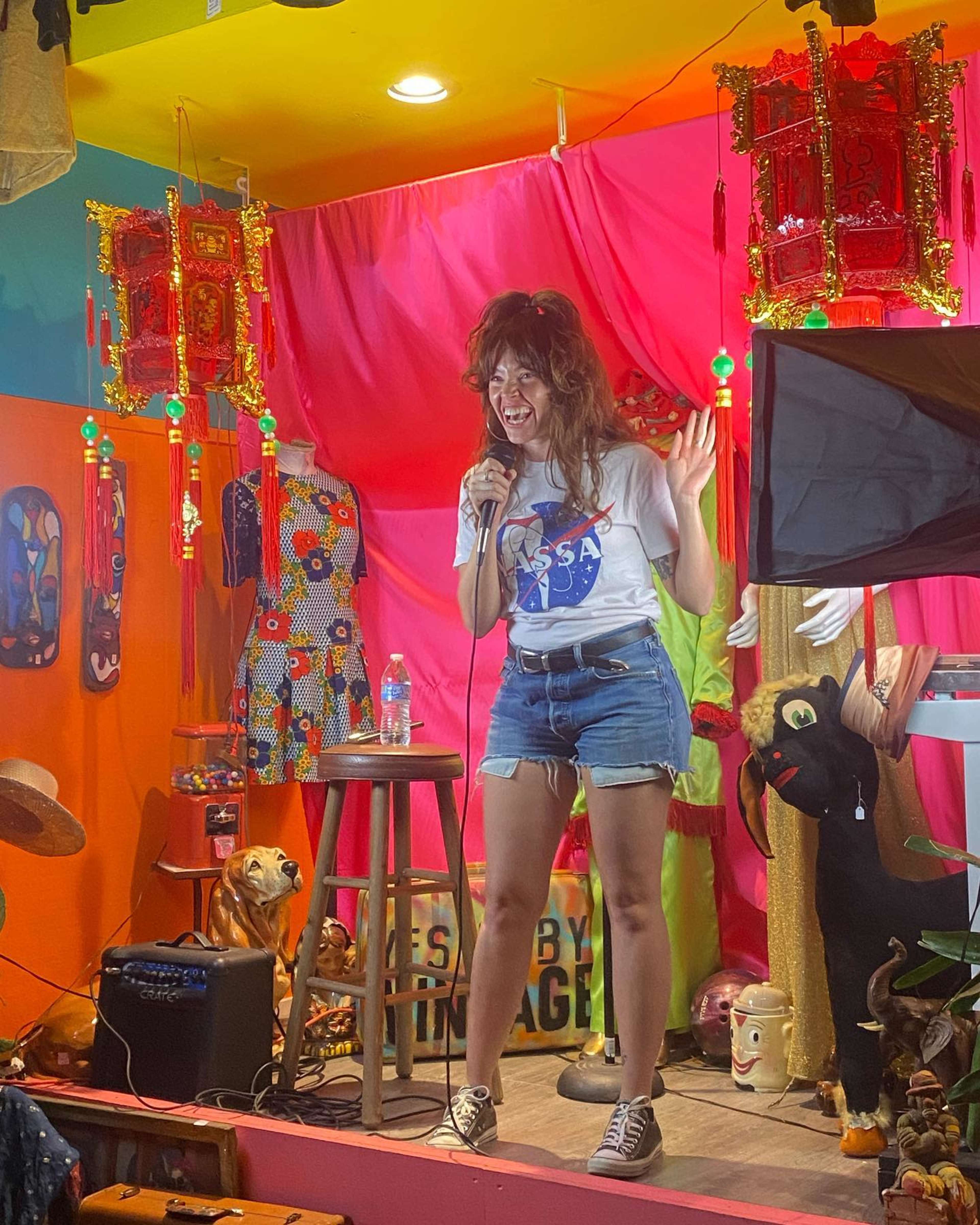 A woman in a NASA-themed t-shirt performs stand-up comedy on a small stage decorated with colorful fabrics and various props.