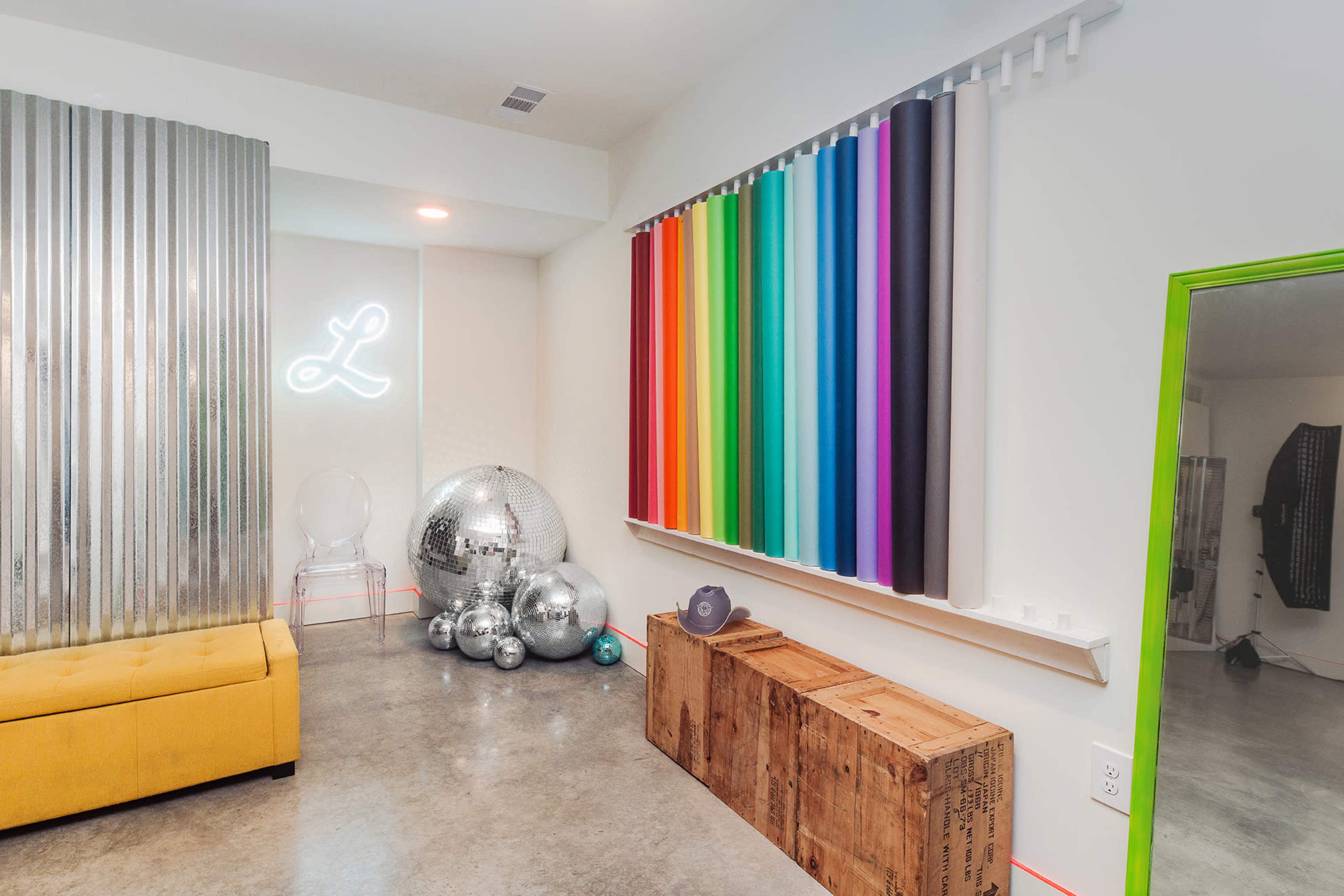 The image shows a brightly colored interior space featuring a yellow sofa, a wall with various colored vertical stripes, and shiny disco balls arranged near a wooden crate.