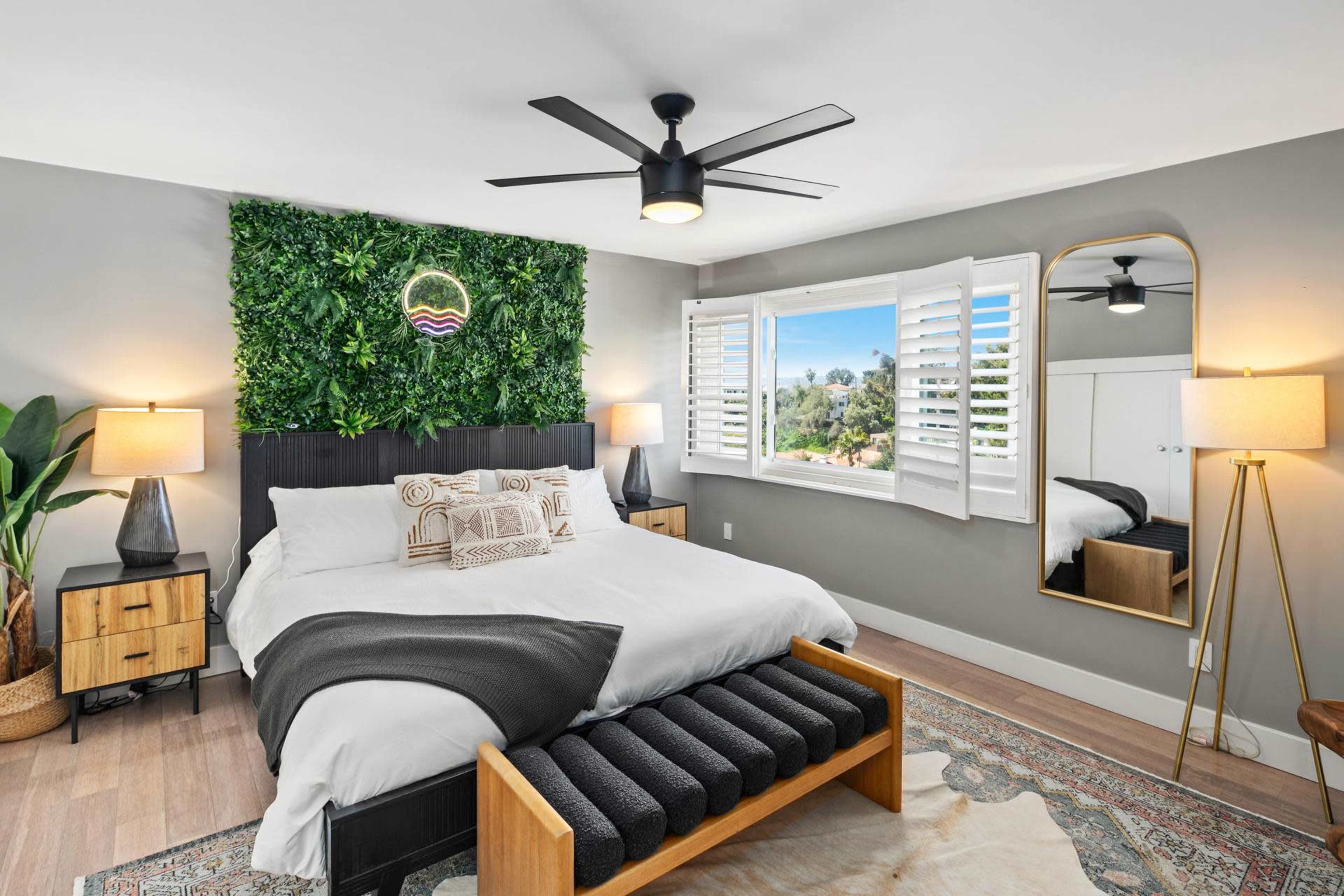 The bedroom features a large bed with a green wall decoration, large windows with shutters, and a mirror beside a standing lamp.