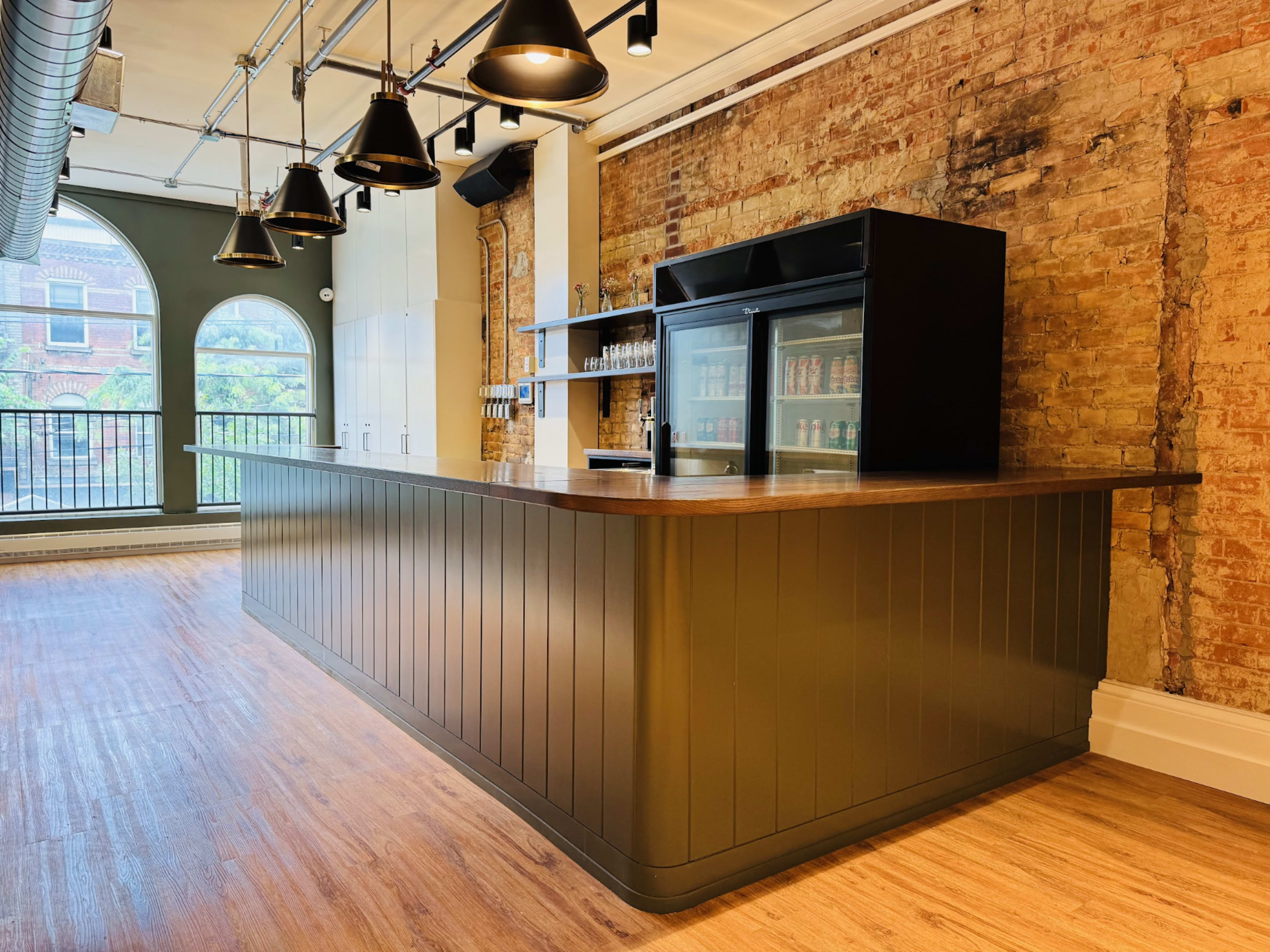 A modern bar counter with dark cabinetry and a glass display fridge is set against a backdrop of exposed brick walls and large windows.