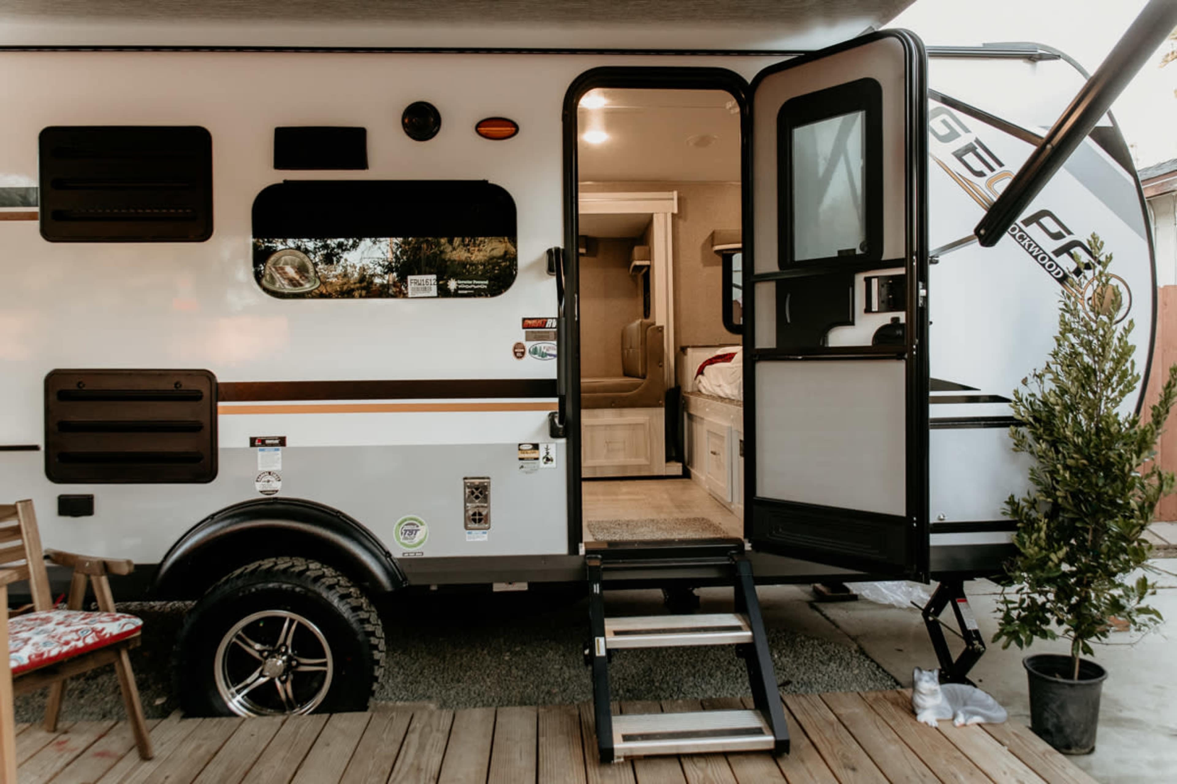A travel trailer is parked outside on a wooden deck, showcasing an entryway with steps leading into the interior.