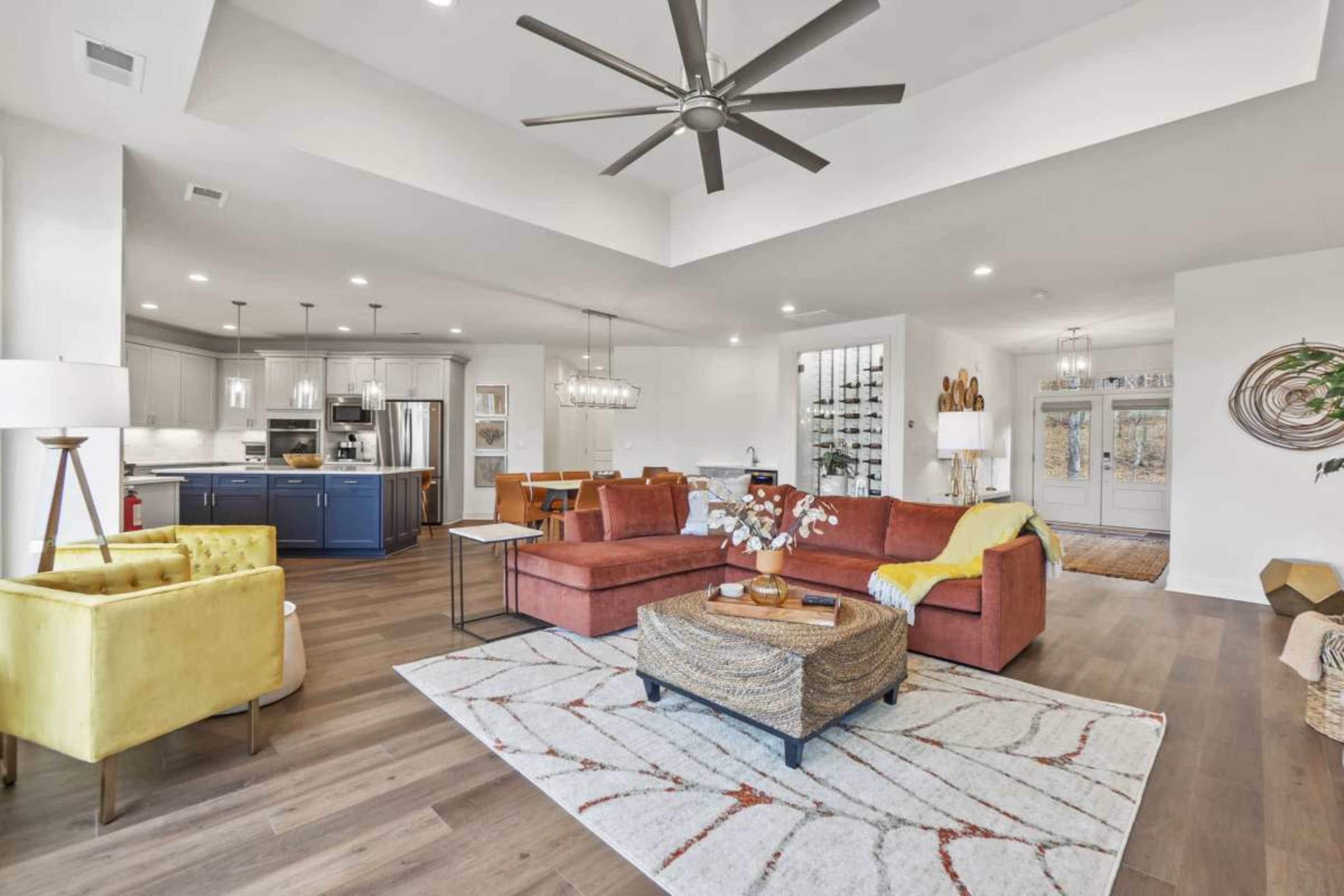 A modern living room features a large sectional sofa, vibrant armchairs, and a textured area rug, with a dining area and kitchen visible in the background.