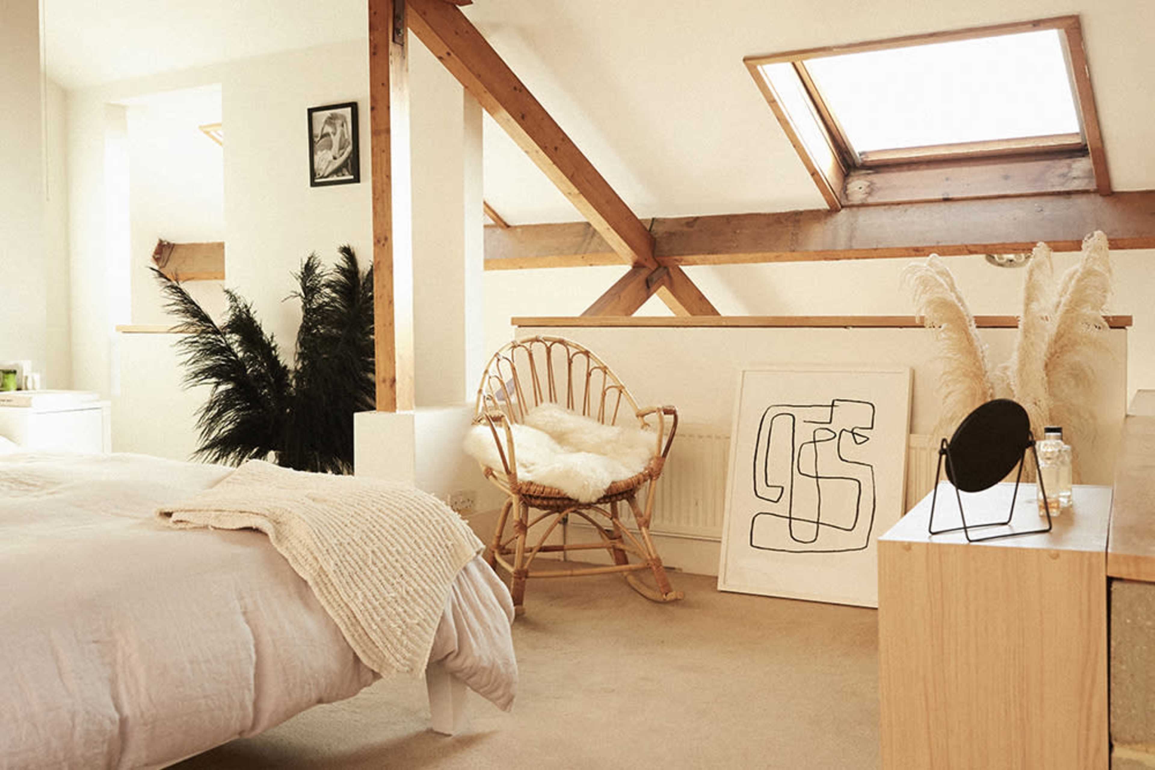 A cozy attic bedroom features a bed, a rattan chair, and a minimalist art piece against white walls and wooden beams.