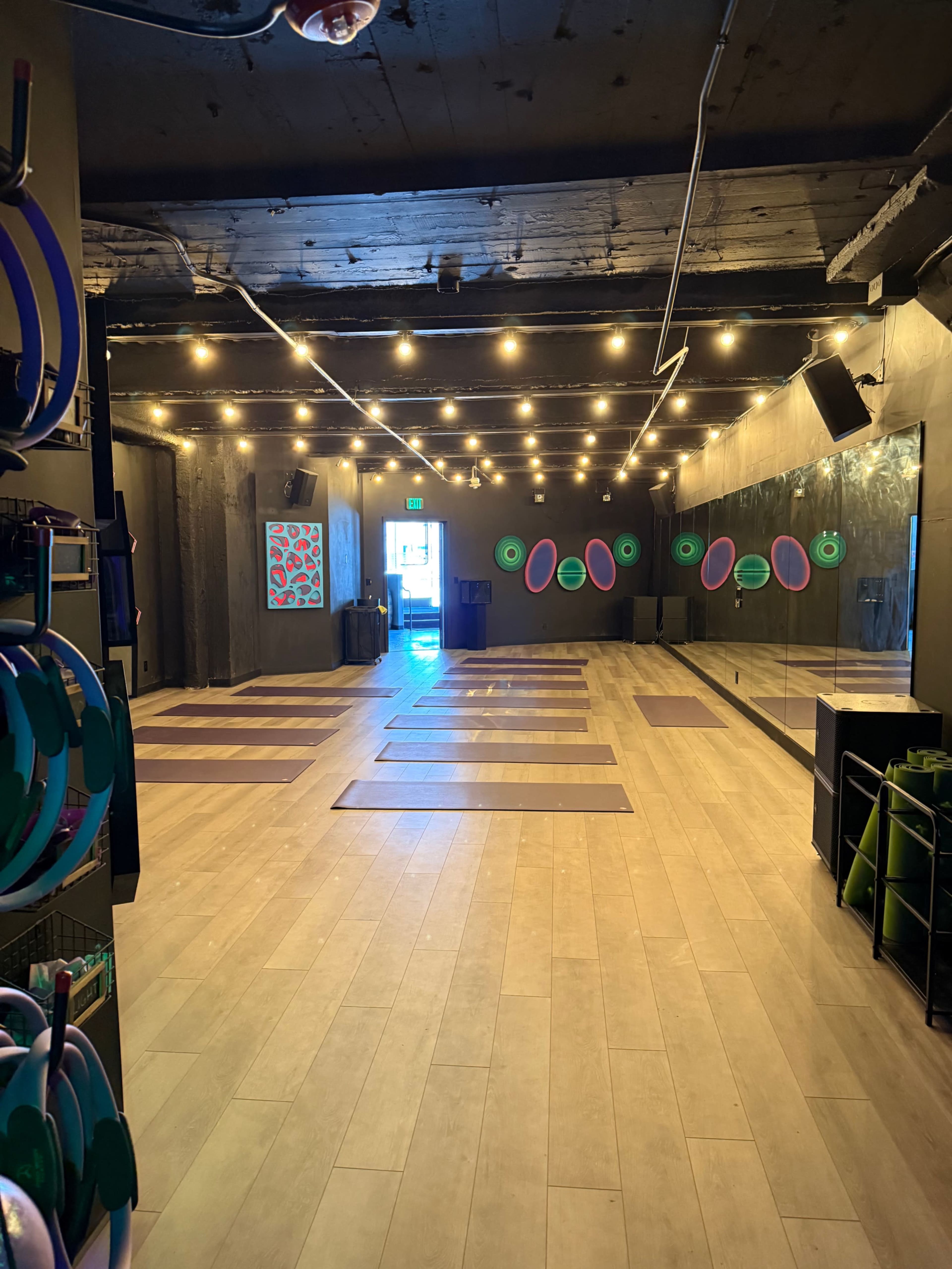 The photo shows an empty yoga studio with several yoga mats placed on a wooden floor, illuminated by string lights and featuring a large mirror along one wall.