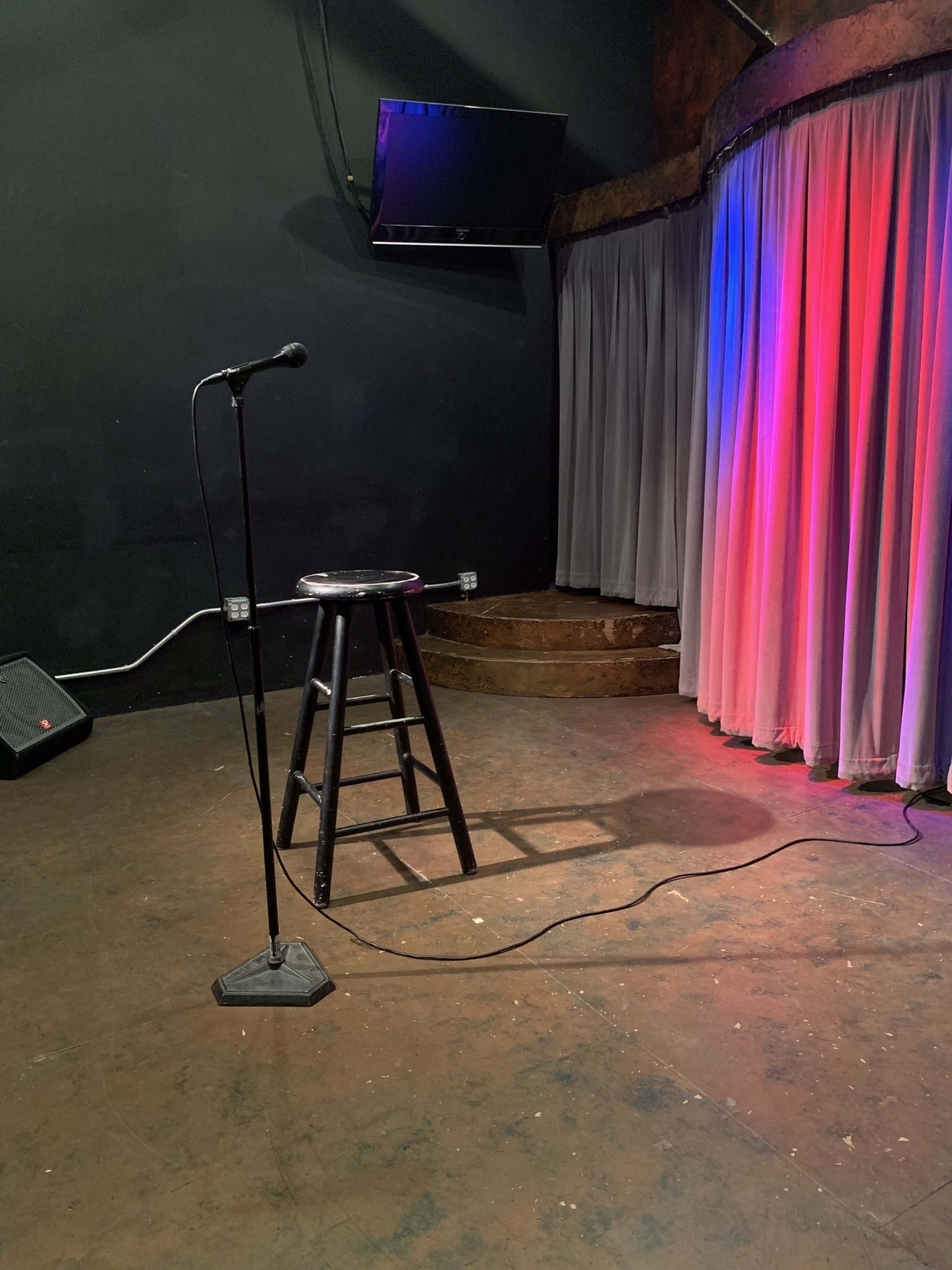 A dimly lit stage with a black microphone stand, a stool, and curtains in red and blue lighting.