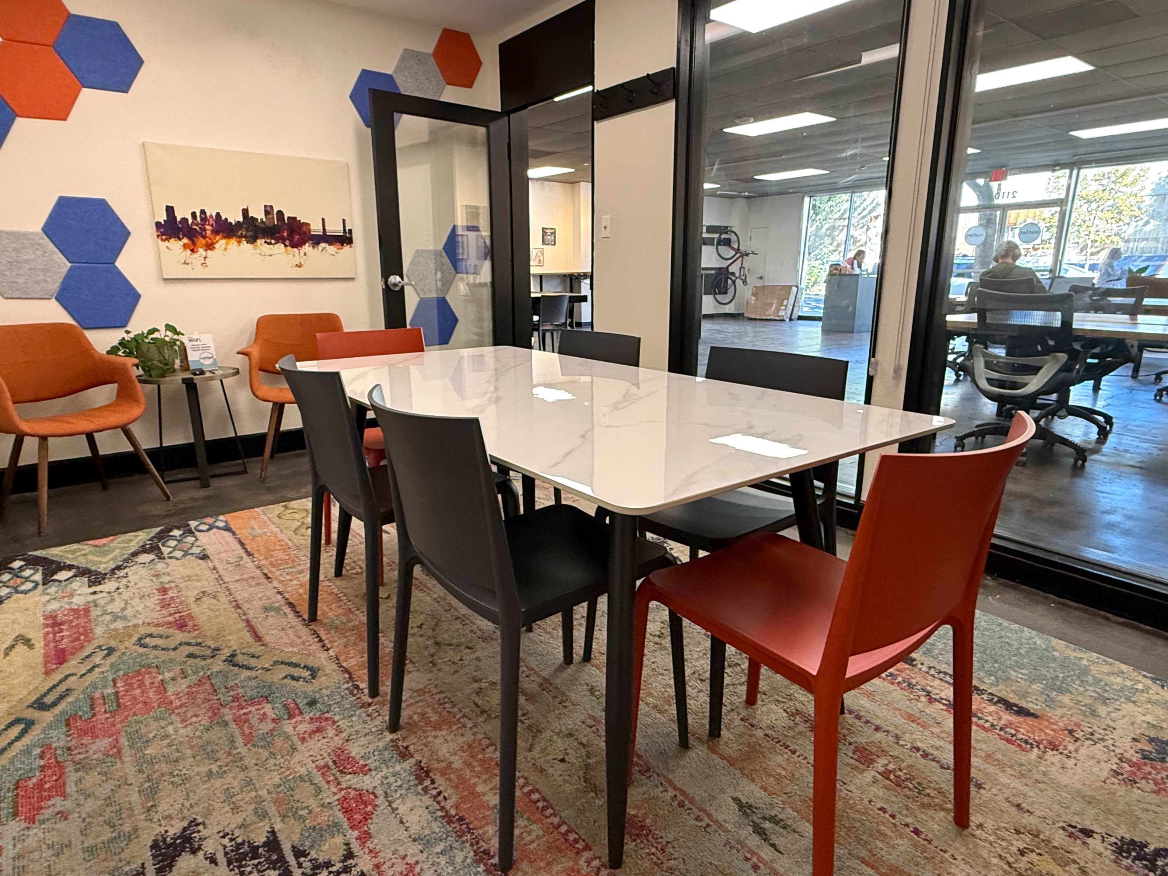 A rectangular white table surrounded by six chairs of various colors sits in a modern office space with hexagonal wall art and a patterned rug.