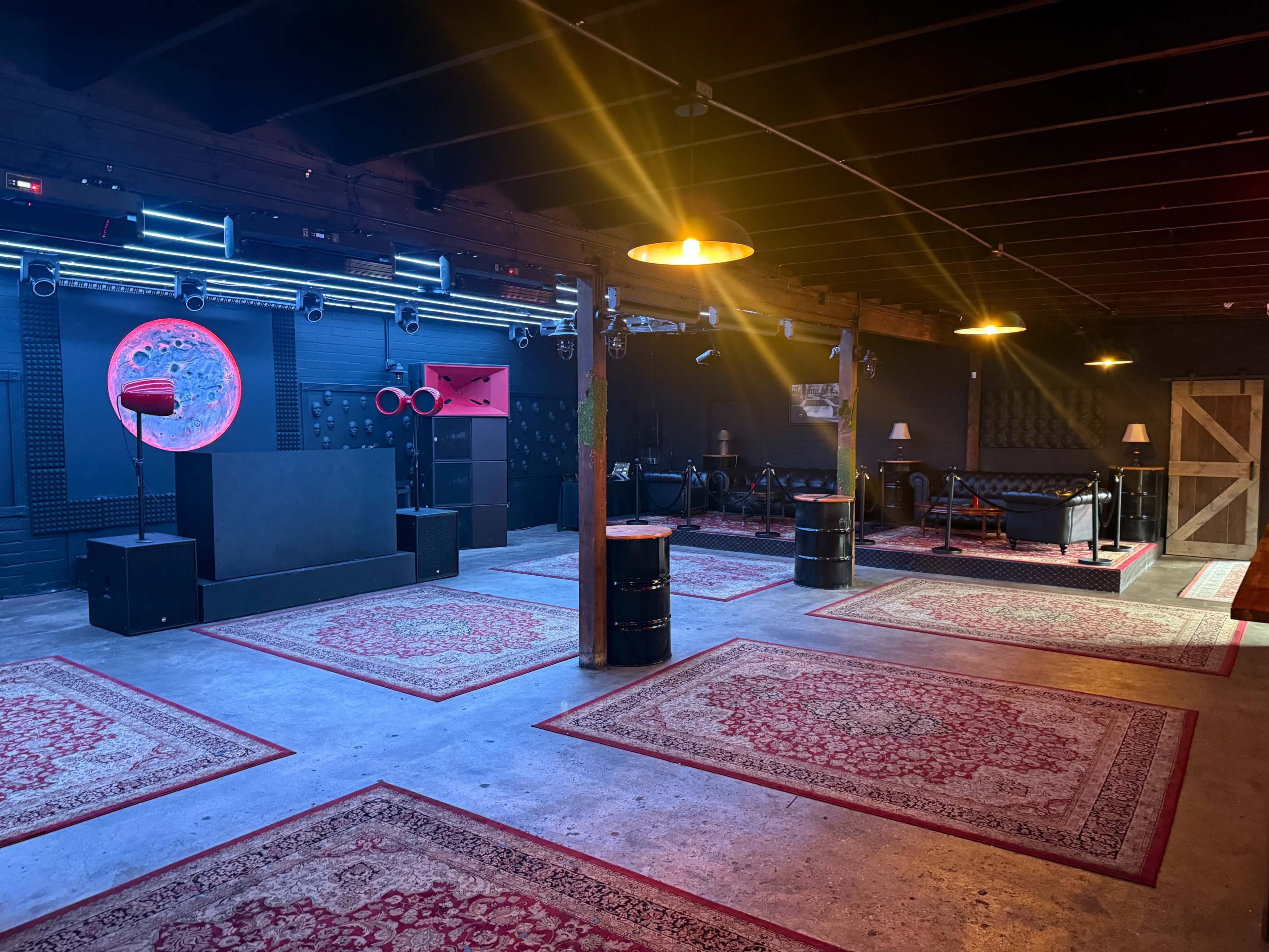 The image shows an empty, dimly lit event space featuring patterned rugs, a stage area, and various lighting fixtures.
