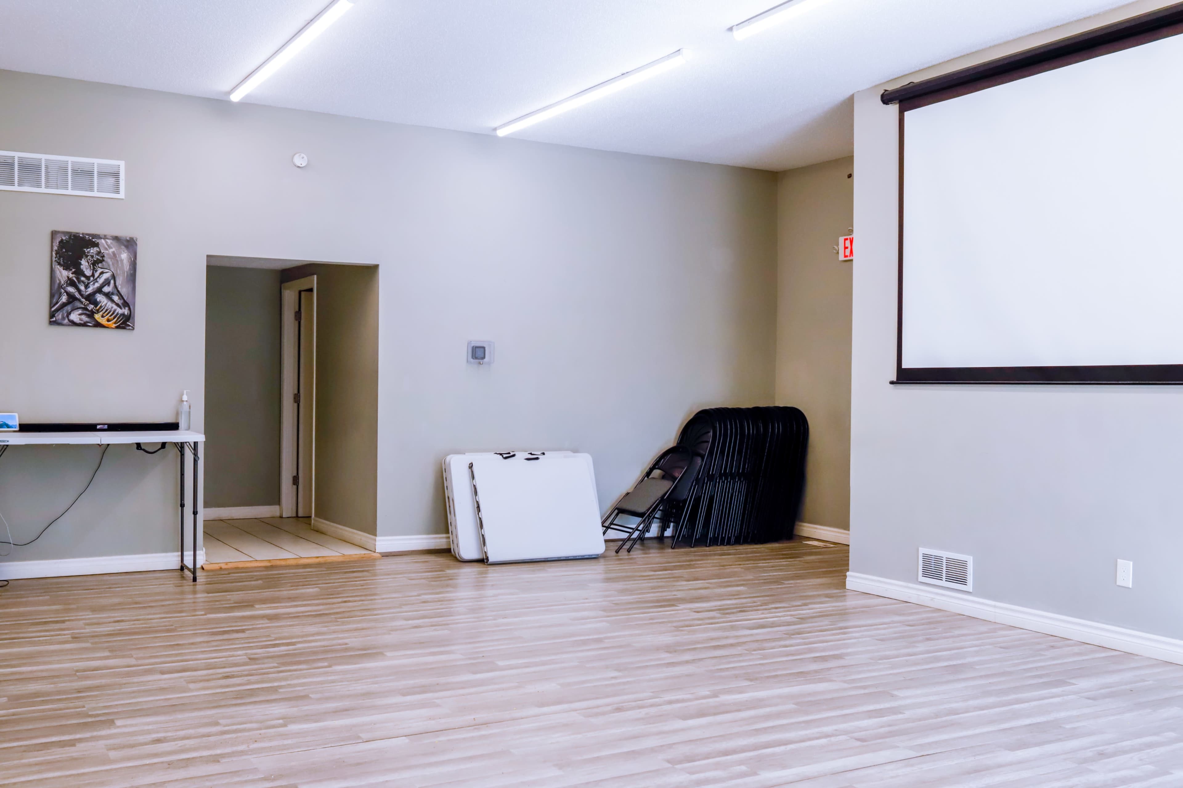 A spacious room with hardwood flooring, featuring a projector screen on one wall, a table with a laptop, and stacked folding chairs in the corner.