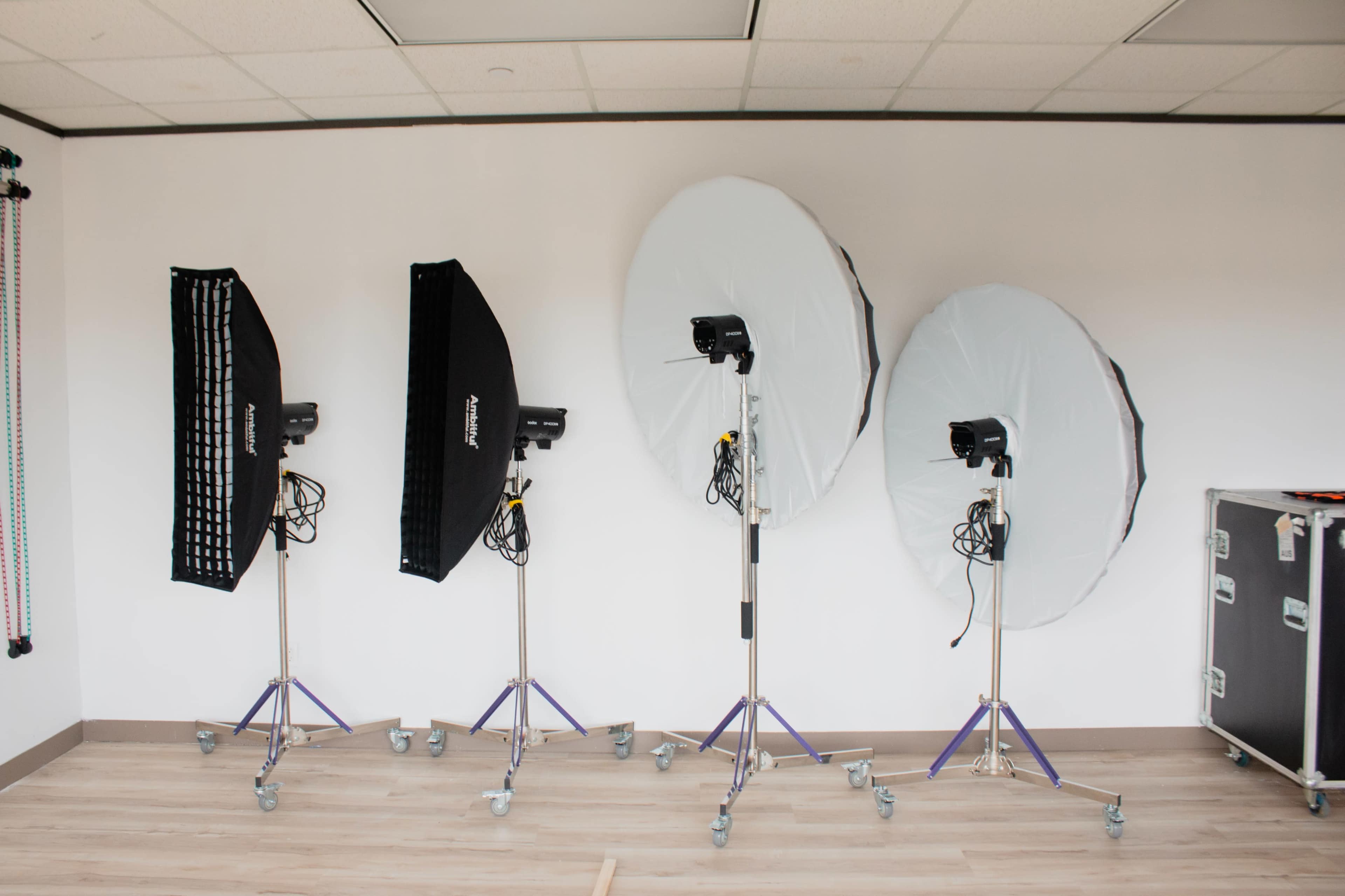 The image shows a photography studio setup with four light stands equipped with softboxes and reflectors against a white wall.