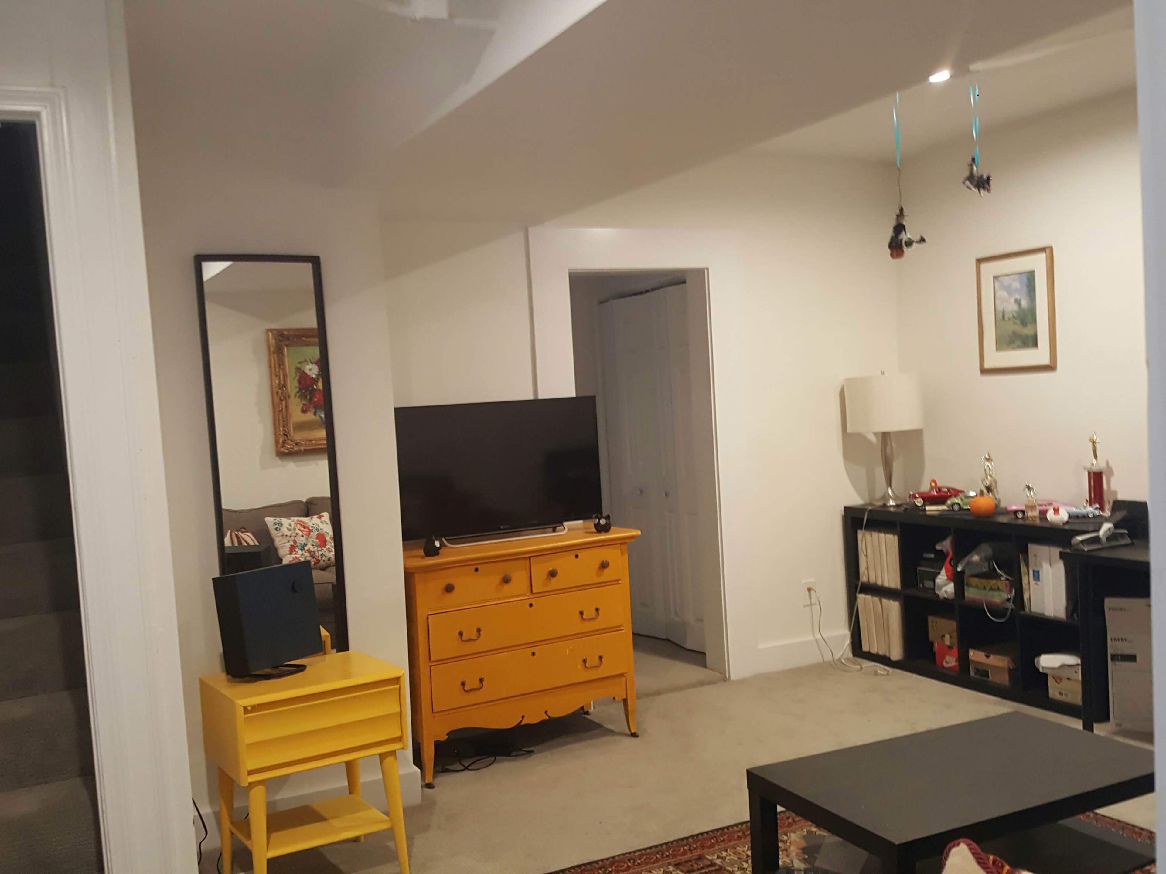 A well-lit living space featuring a yellow dresser, a television on a stand, a bookshelf, and a small yellow table.