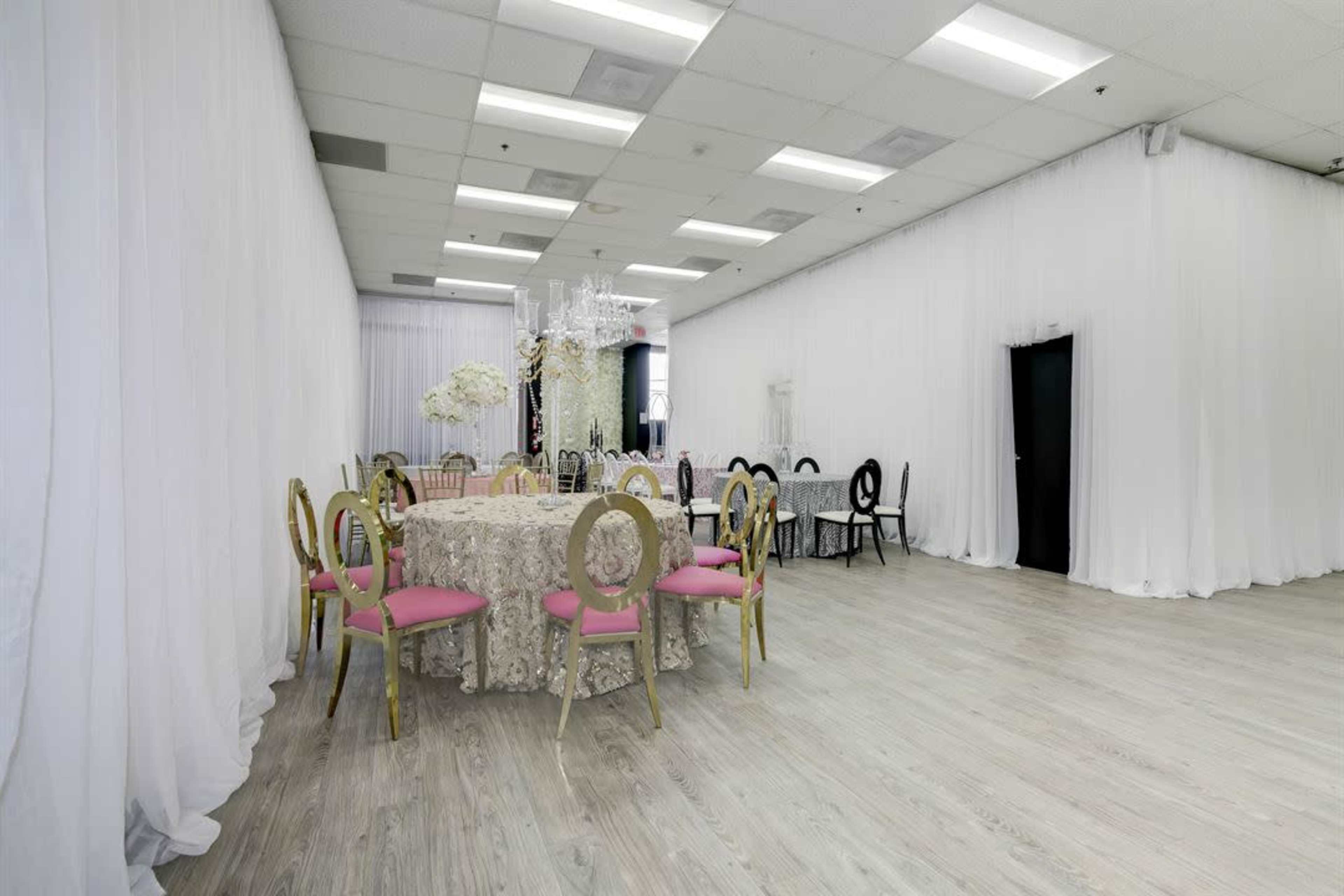 A spacious event hall features a round table with a lace tablecloth and pink chairs, surrounded by draped white fabric.