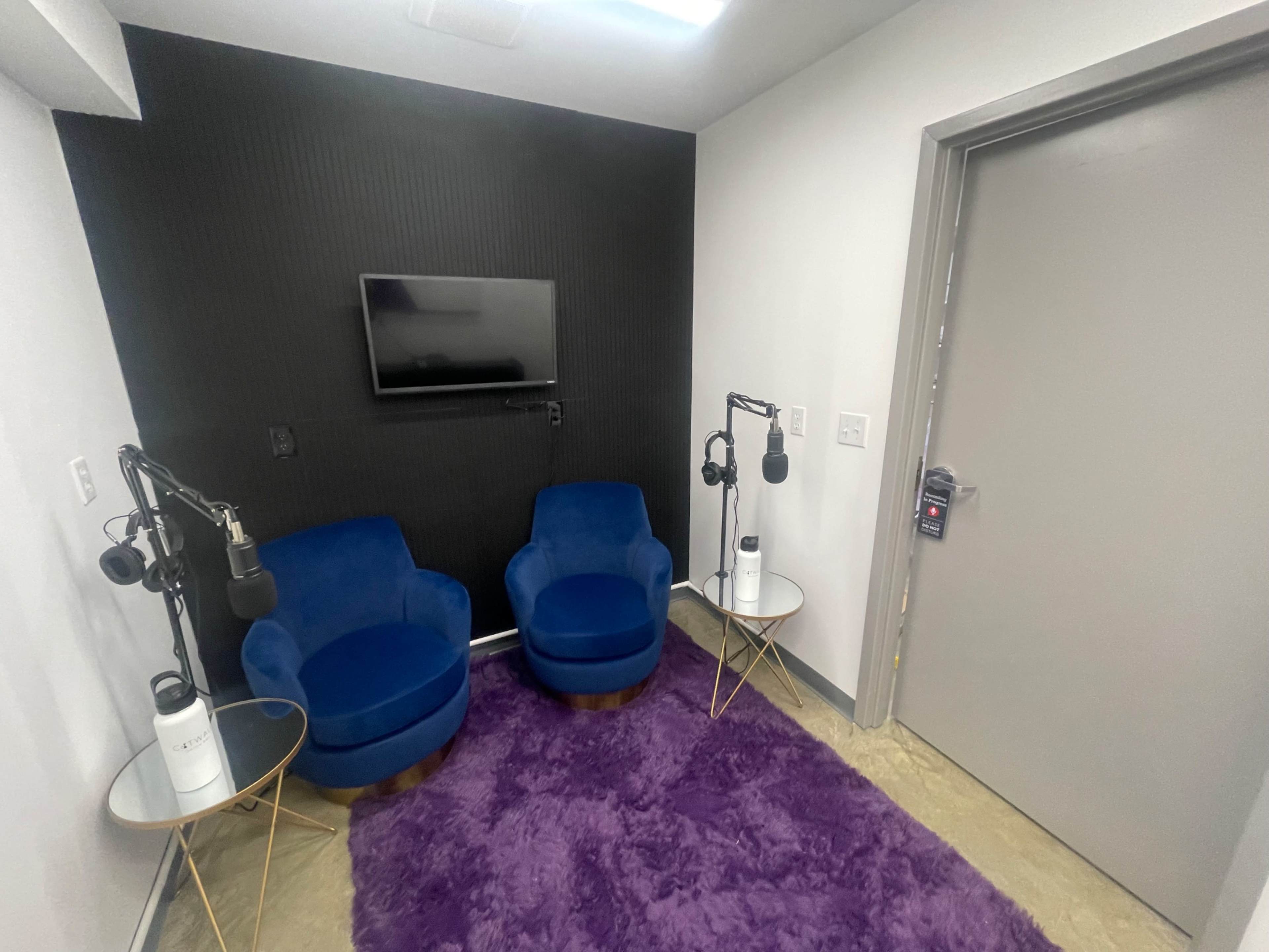 The image shows a small recording room with two blue chairs, a purple rug, a television on the wall, and two microphones mounted on stands.