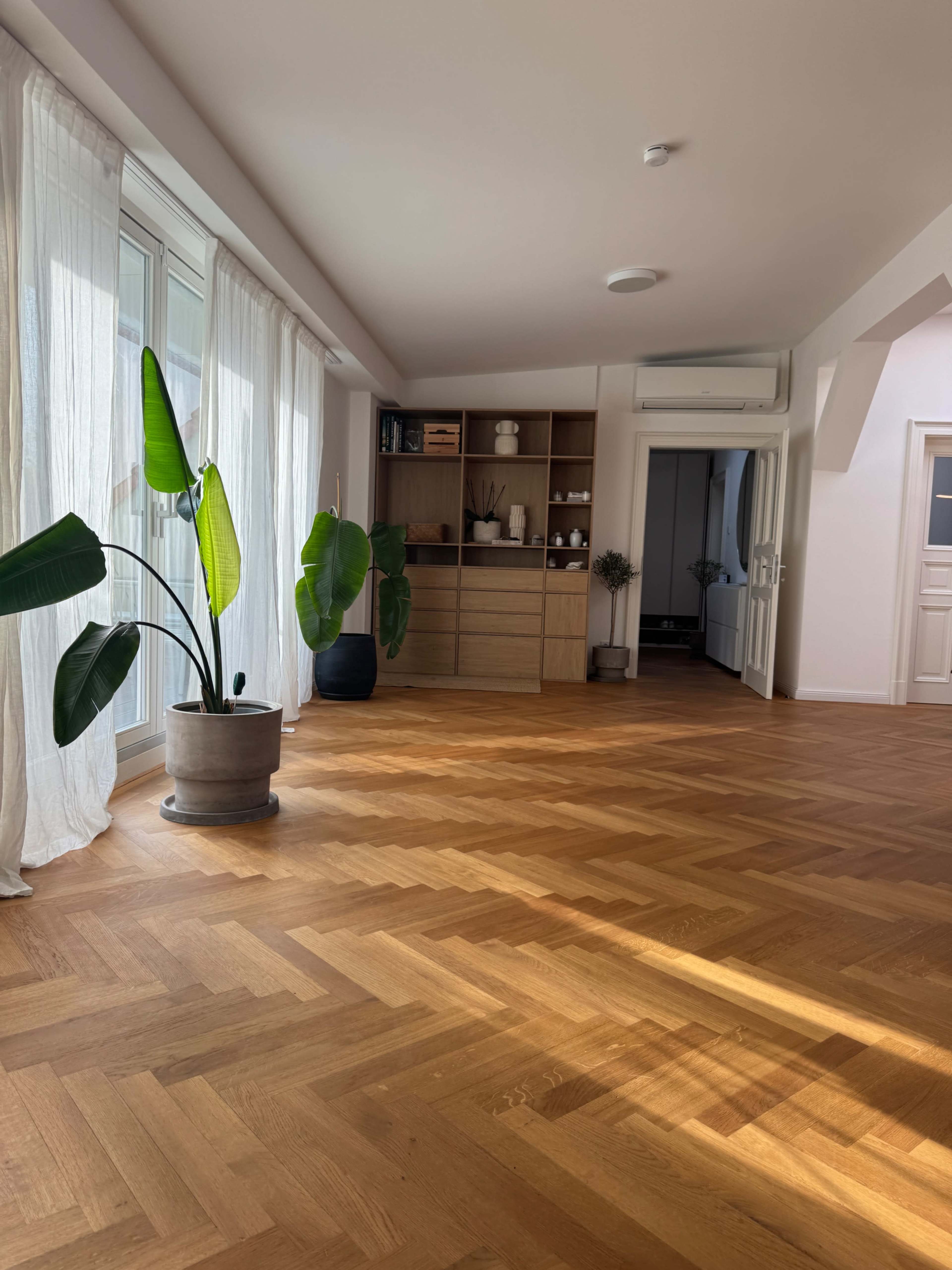 The image shows a spacious, bright room with a herringbone wood floor, large windows covered with sheer curtains, and a shelf filled with various items against one wall.