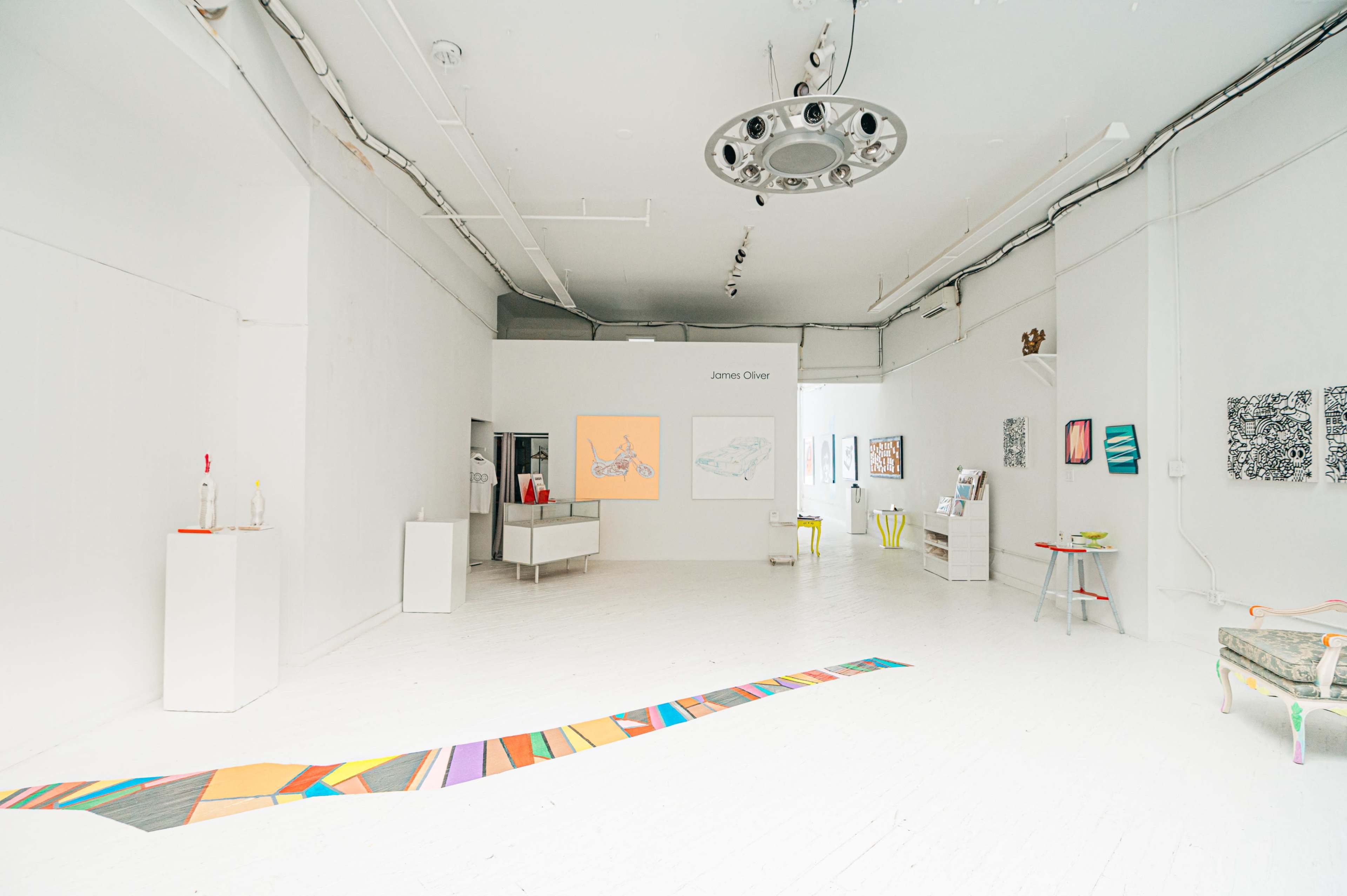 A spacious, well-lit art gallery with various colorful artworks displayed on the white walls and a patterned strip running across the floor.