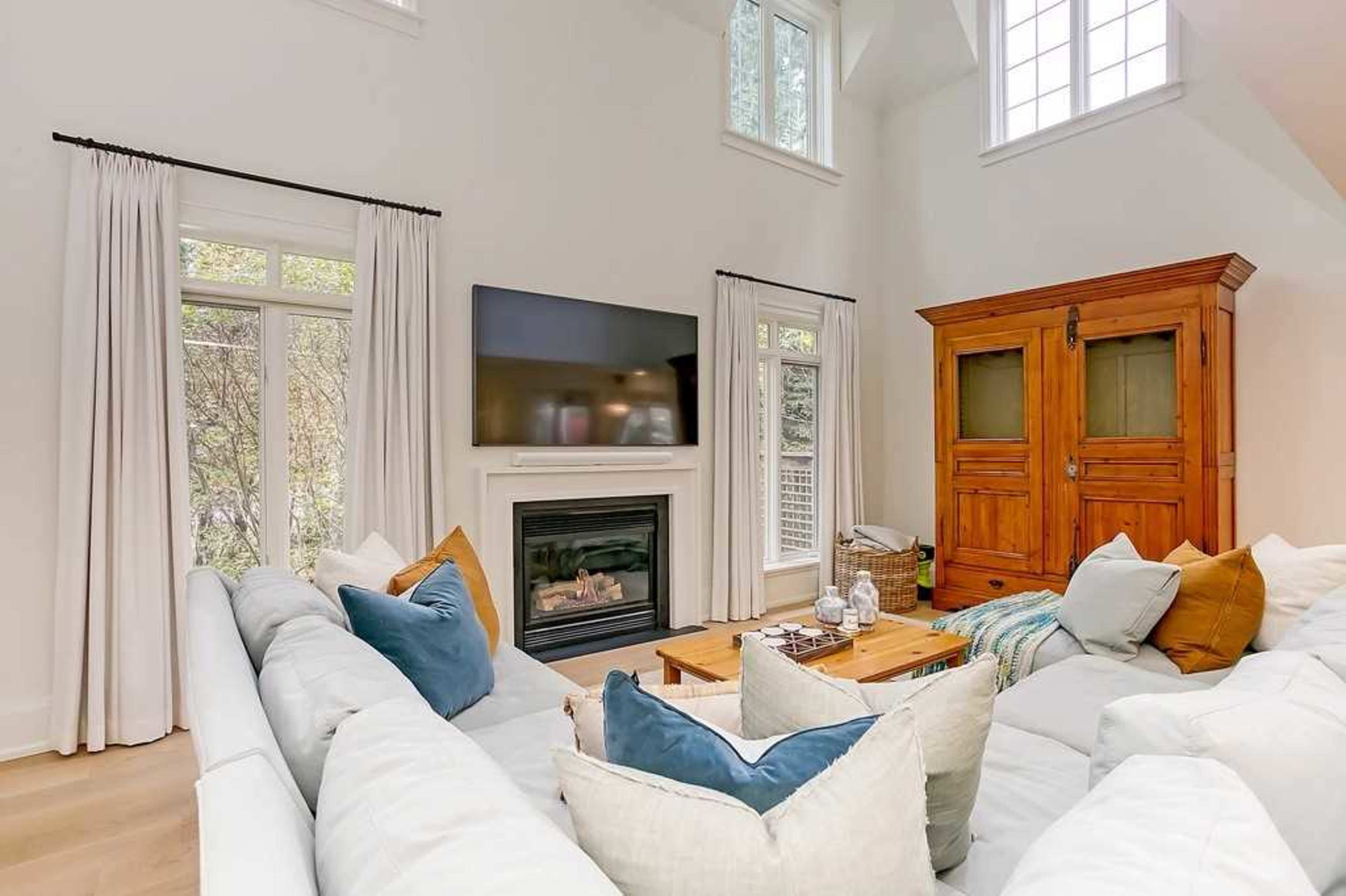 The image shows a modern living room with a large sectional sofa, a fireplace beneath a wall-mounted television, and an antique wooden cabinet.