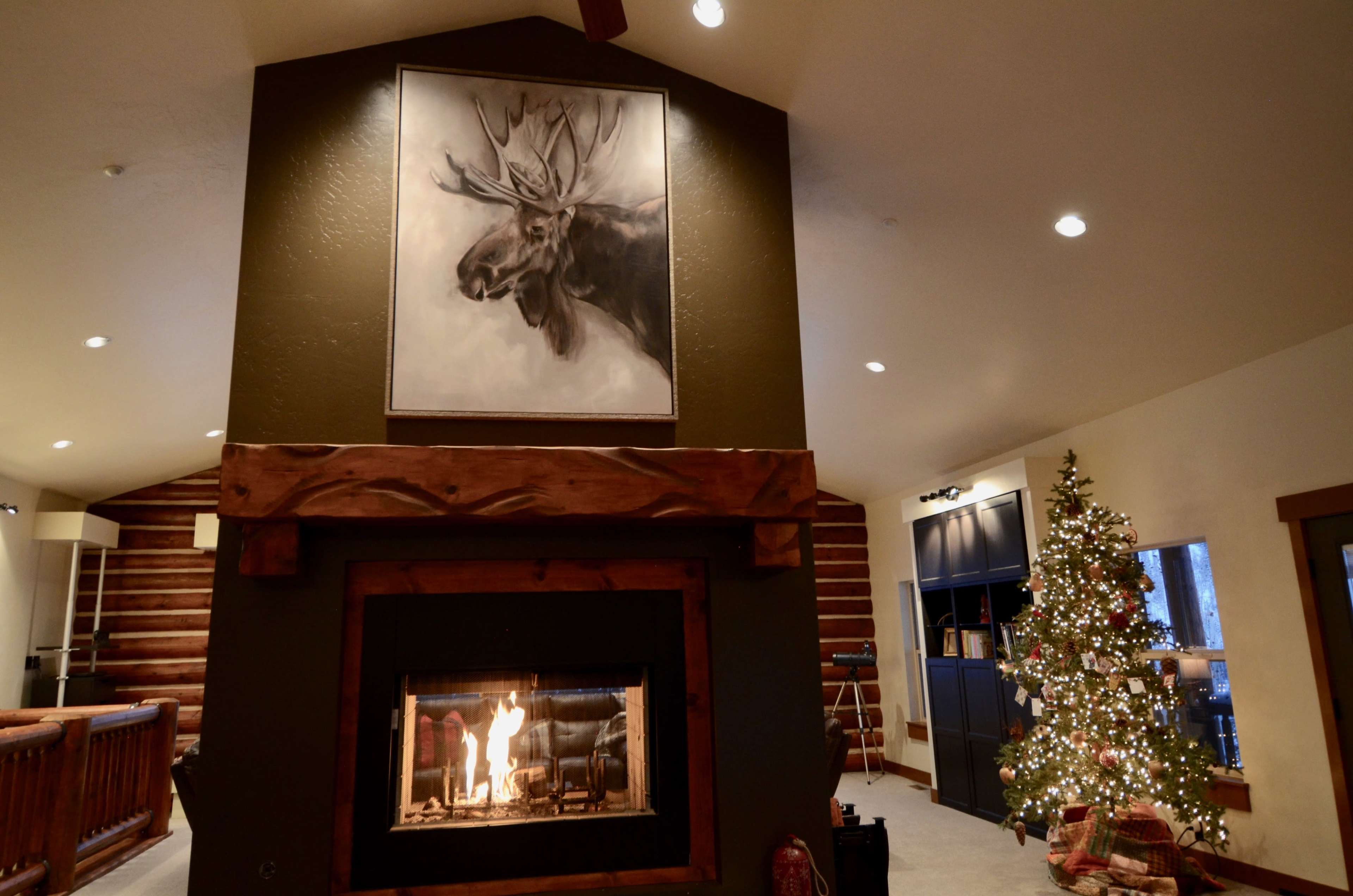 A fireplace is centered on a wall with a moose painting above it, and a Christmas tree stands in the corner of a cozy room.