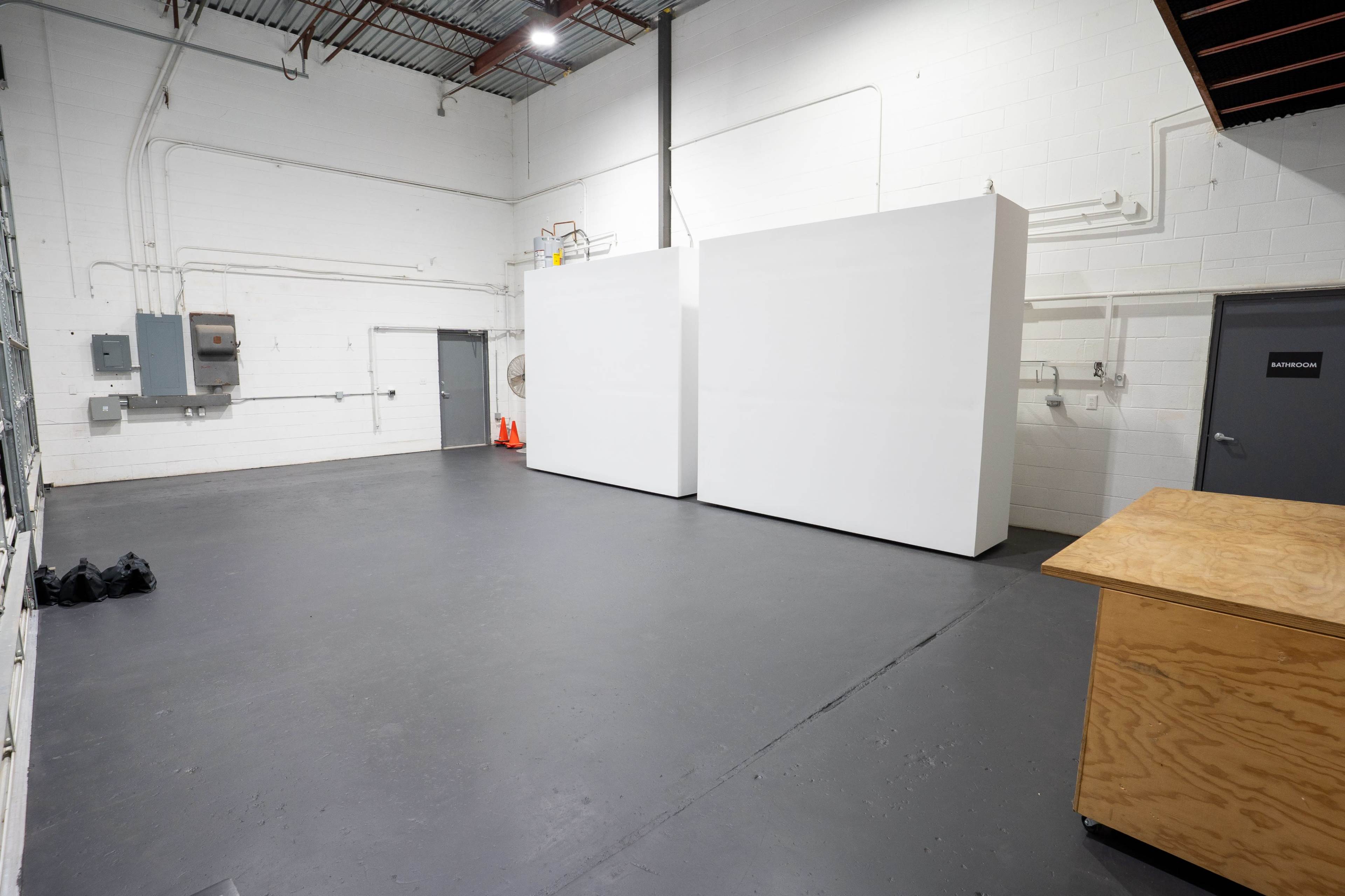 The image shows a spacious, empty room with two large white partitions, a gray door, and a wooden table in the corner.