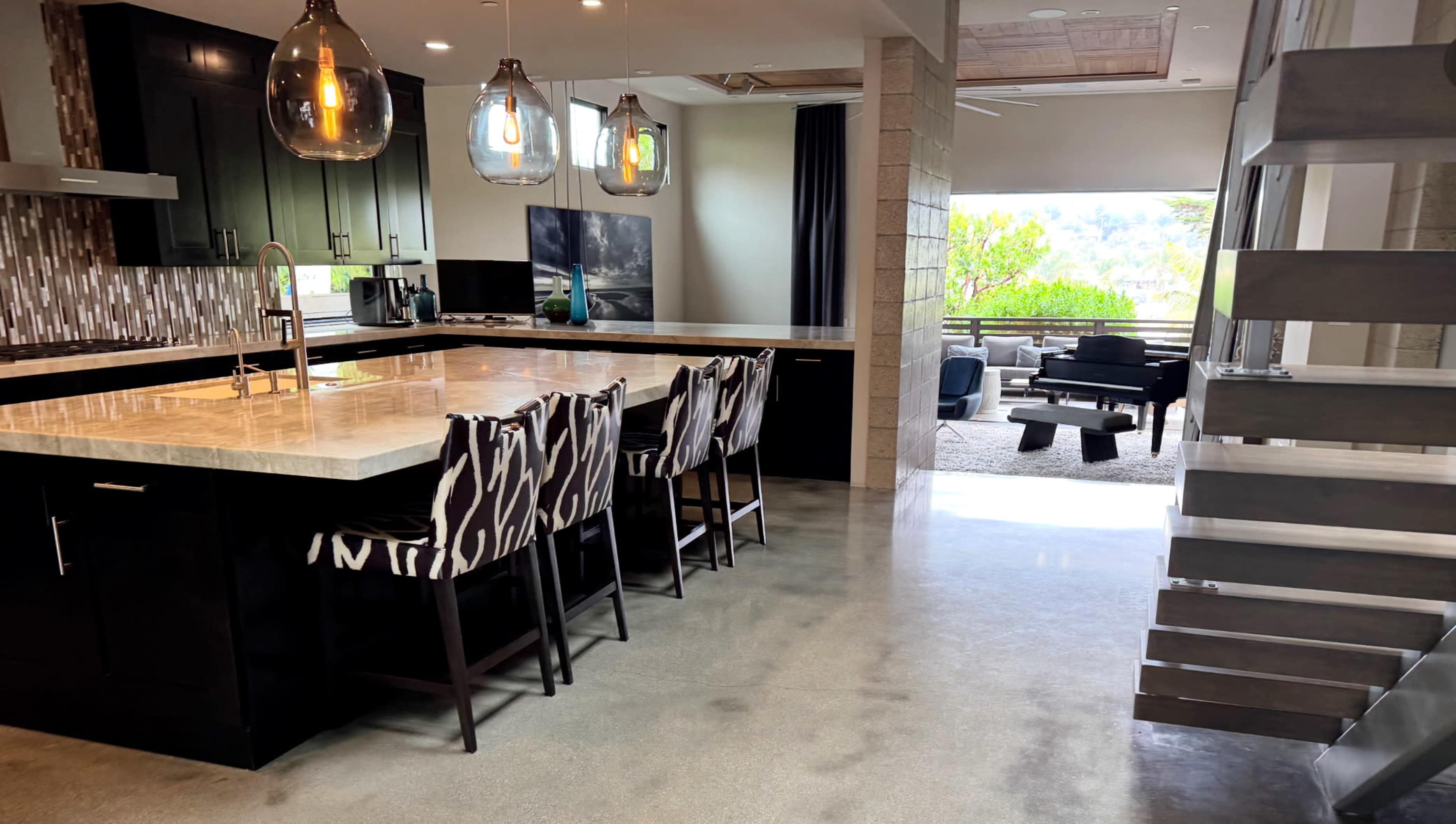 The image shows a modern kitchen with a large island and bar seating, leading to a living area with a piano and large windows overlooking greenery.