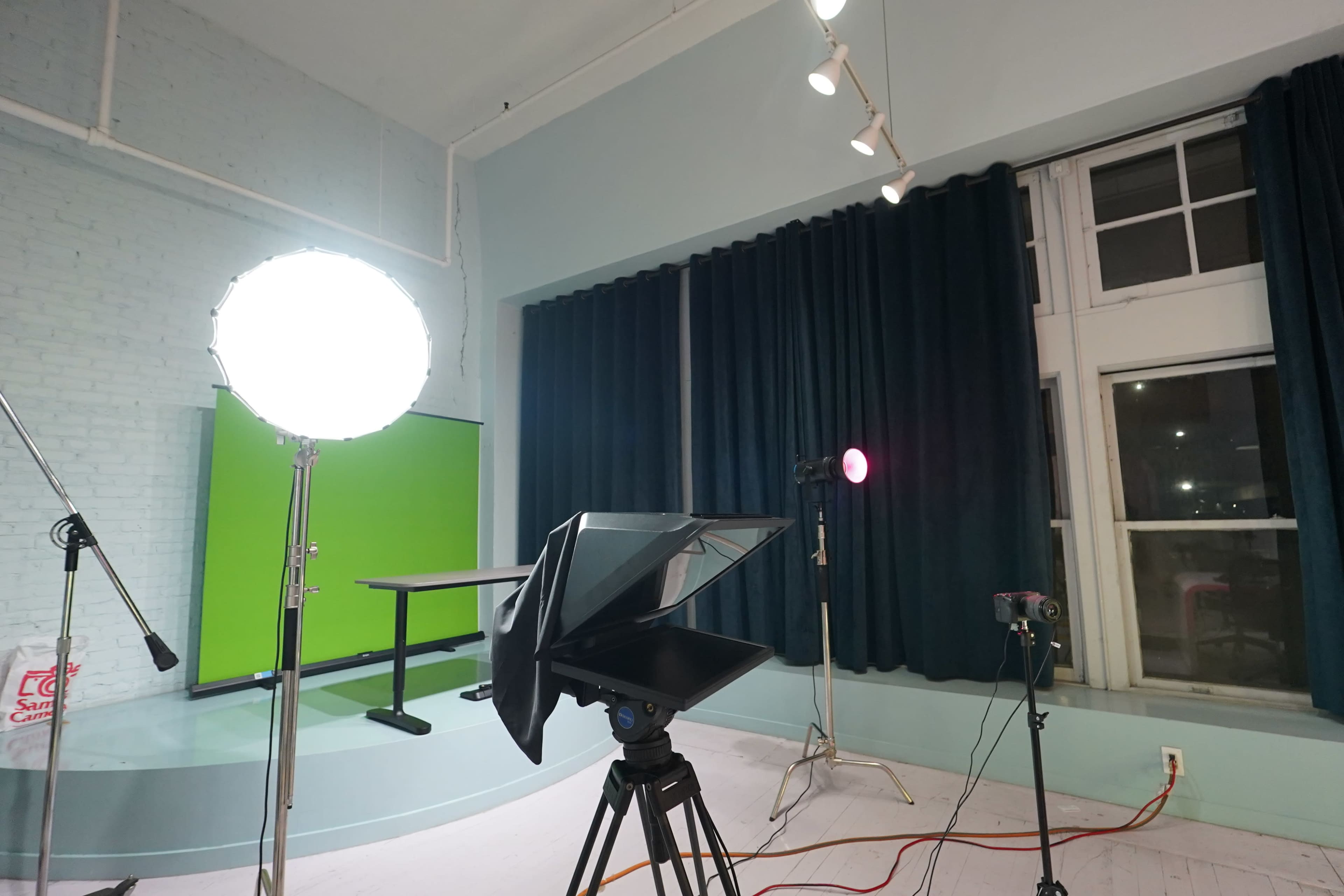 The image shows a video production setup with a teleprompter in the foreground, a bright light source to the left, and a green backdrop on the wall.