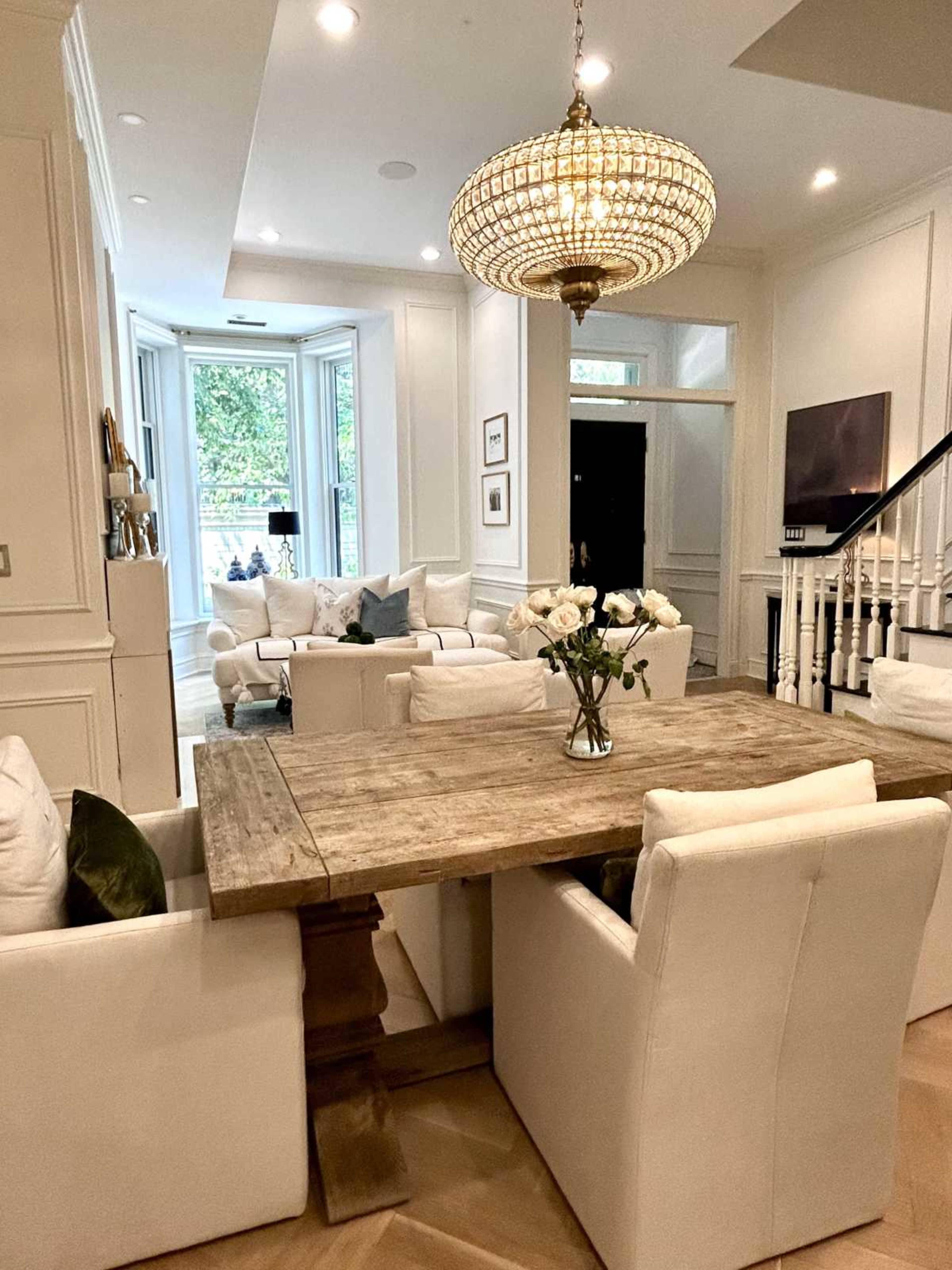 The image features a stylish dining area with a wooden table surrounded by white upholstered chairs, leading into a well-lit living space that includes a chandelier and modern furnishings.
