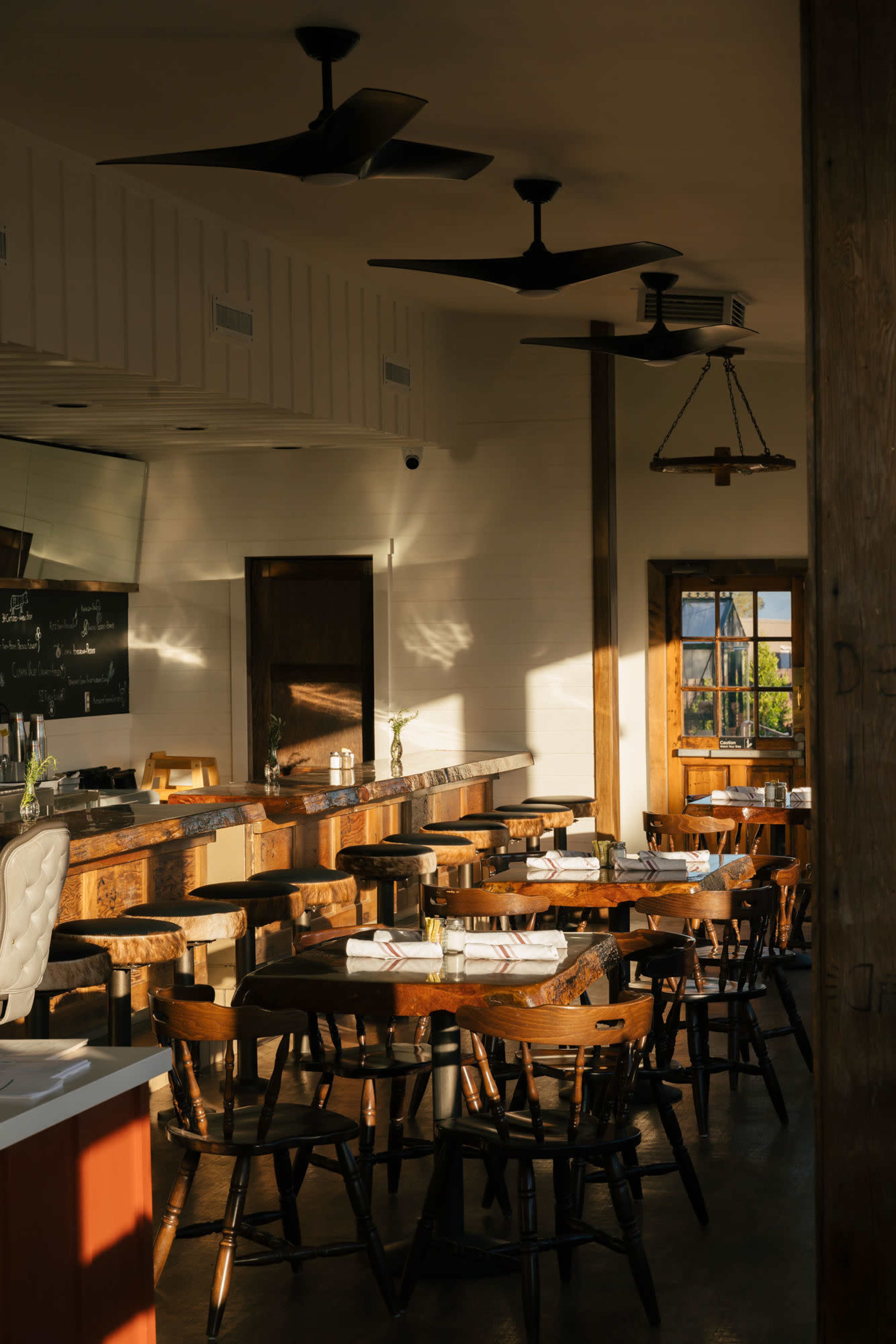 A rustic restaurant interior with wooden tables and chairs, a long bar, and large windows allowing natural light to enter.