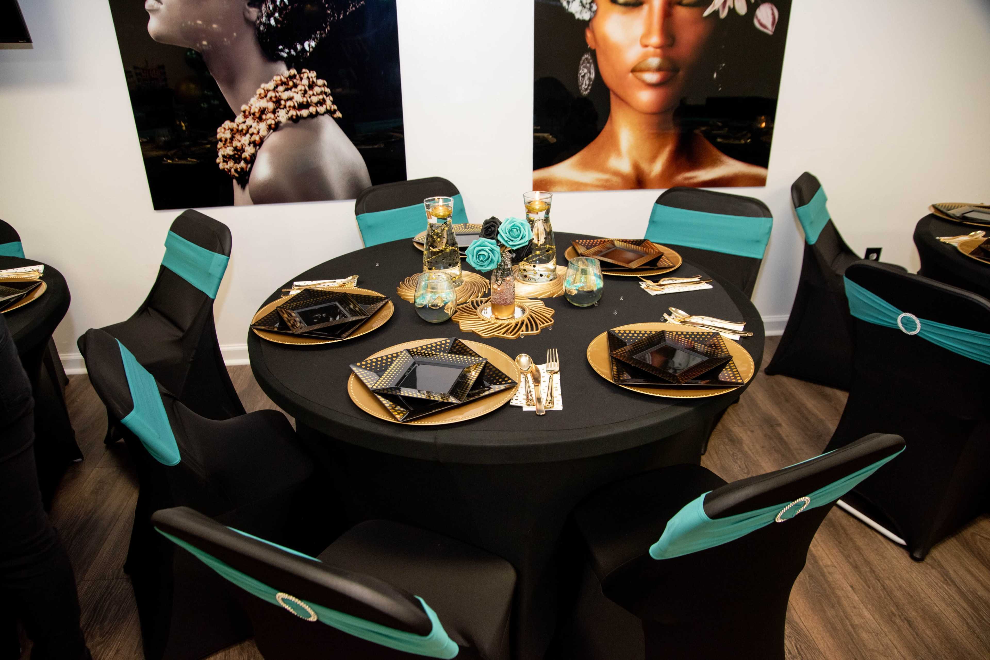A round black dining table elegantly set with gold and black plates, crystal glasses, and turquoise accents, surrounded by chairs covered in black with turquoise sashes.