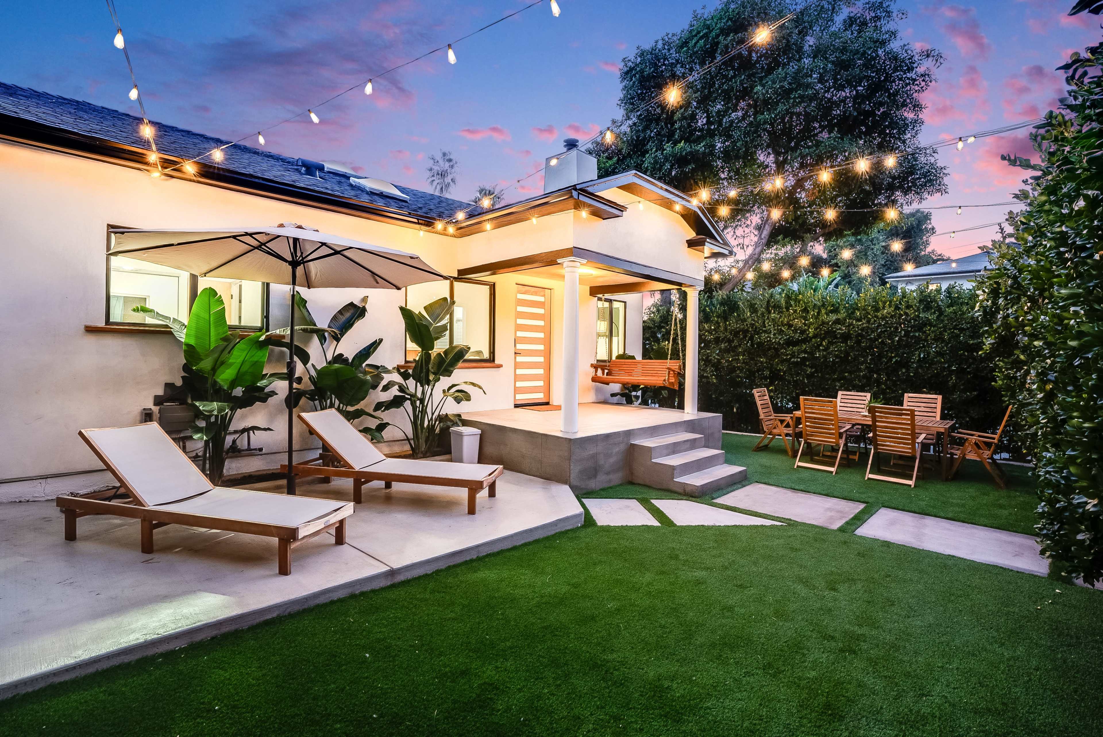 A modern backyard features lounge chairs, an umbrella, and a dining area with wooden chairs, illuminated by string lights at dusk.