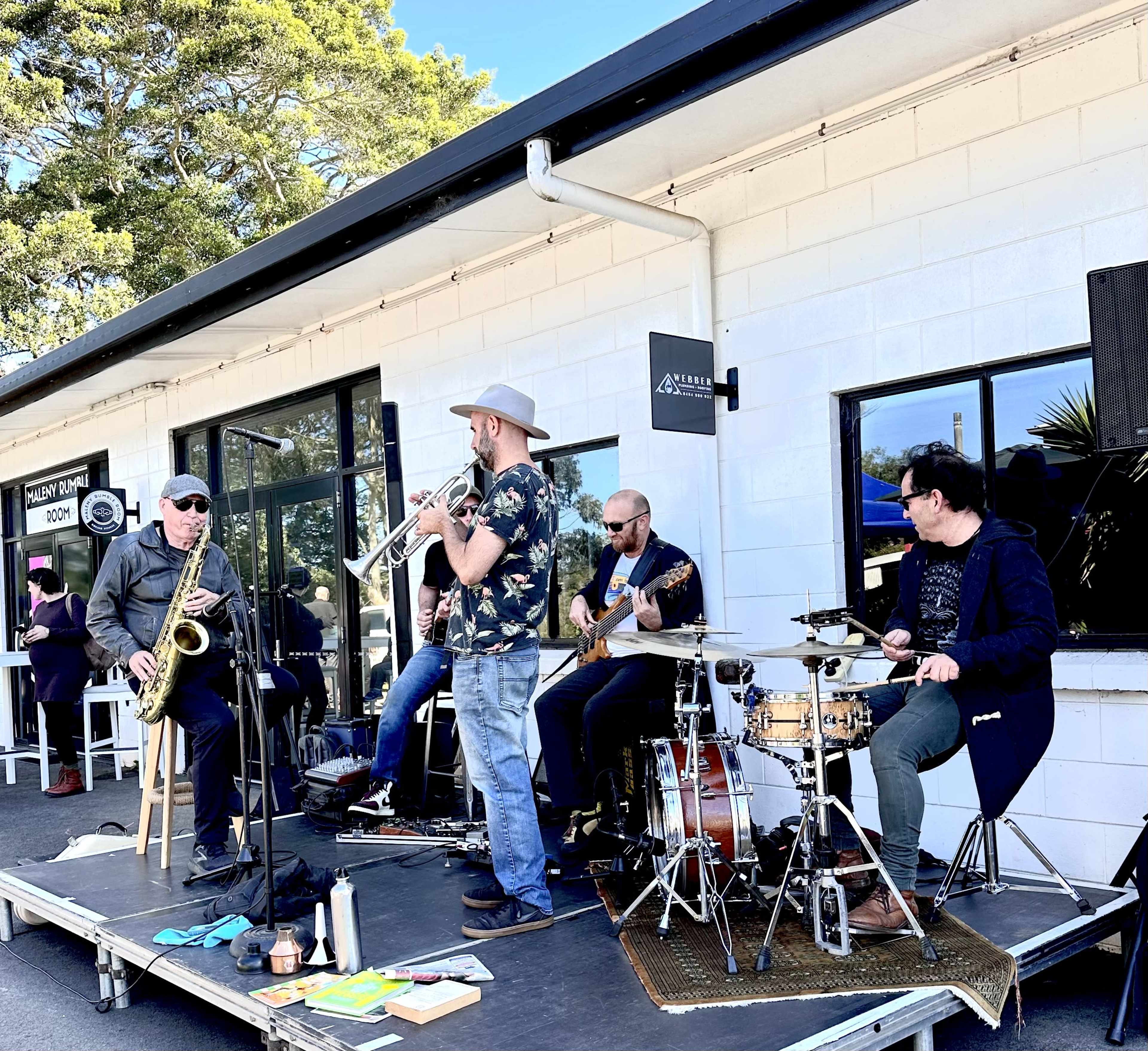 A band performs on a stage outside a building, featuring a saxophonist, a trumpeter, and other musicians playing drums and guitar.