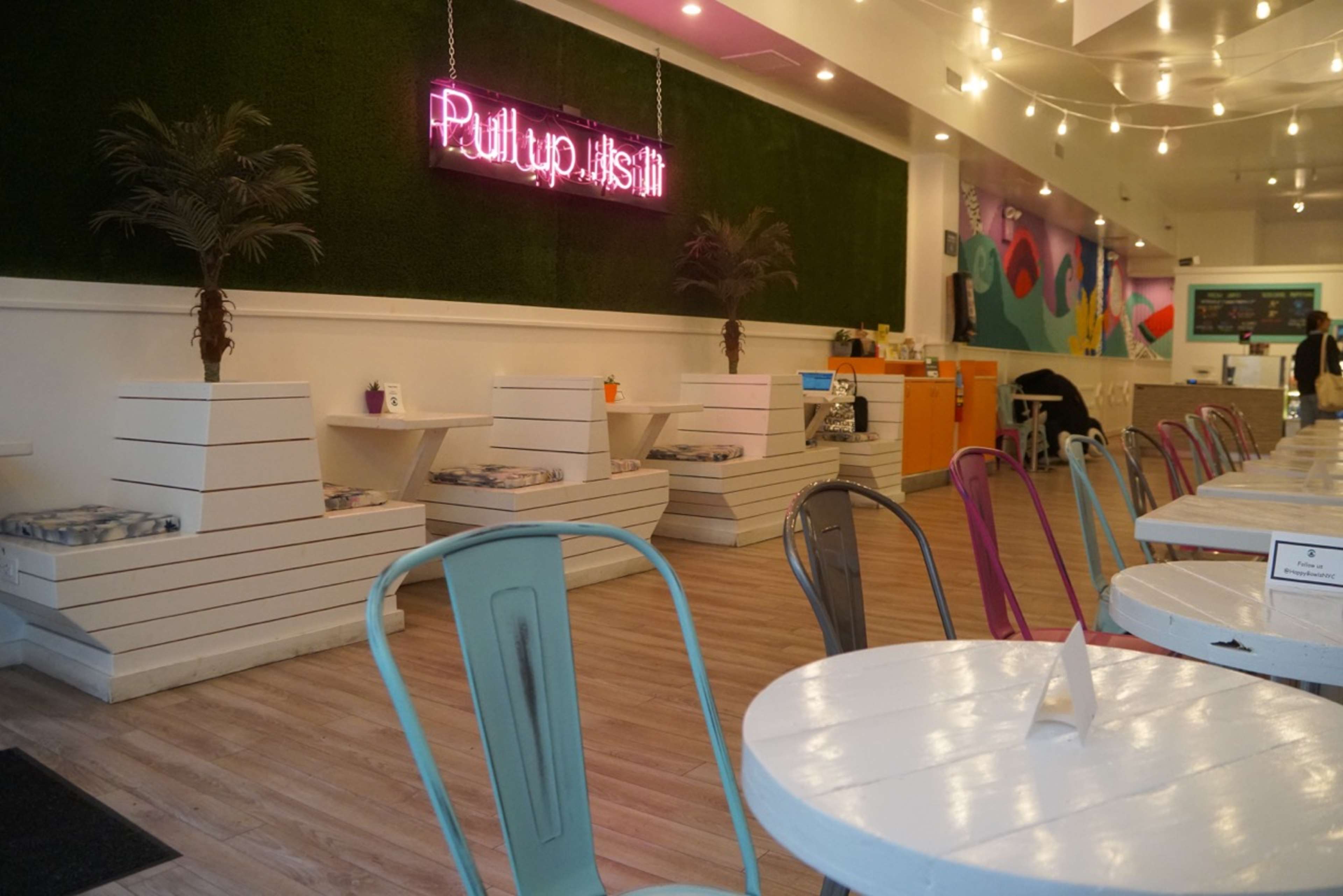 The image shows a bright, modern café interior with wooden floors, white tables, and colorful chairs arranged in rows, accompanied by a neon sign on a green wall.