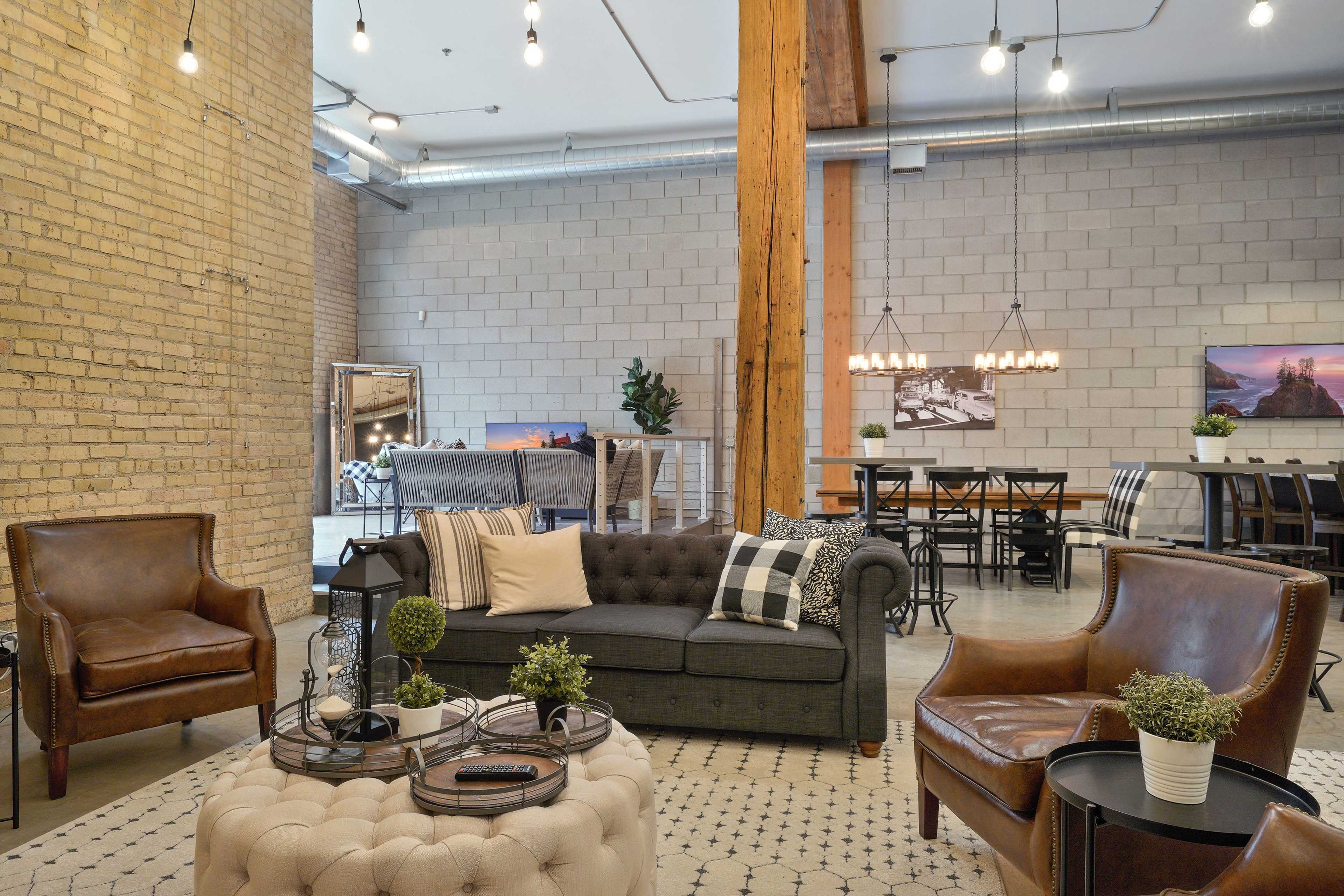 A spacious, modern living area with a mix of seating options, including leather armchairs and a chesterfield sofa, surrounded by exposed brick walls and industrial-style lighting.
