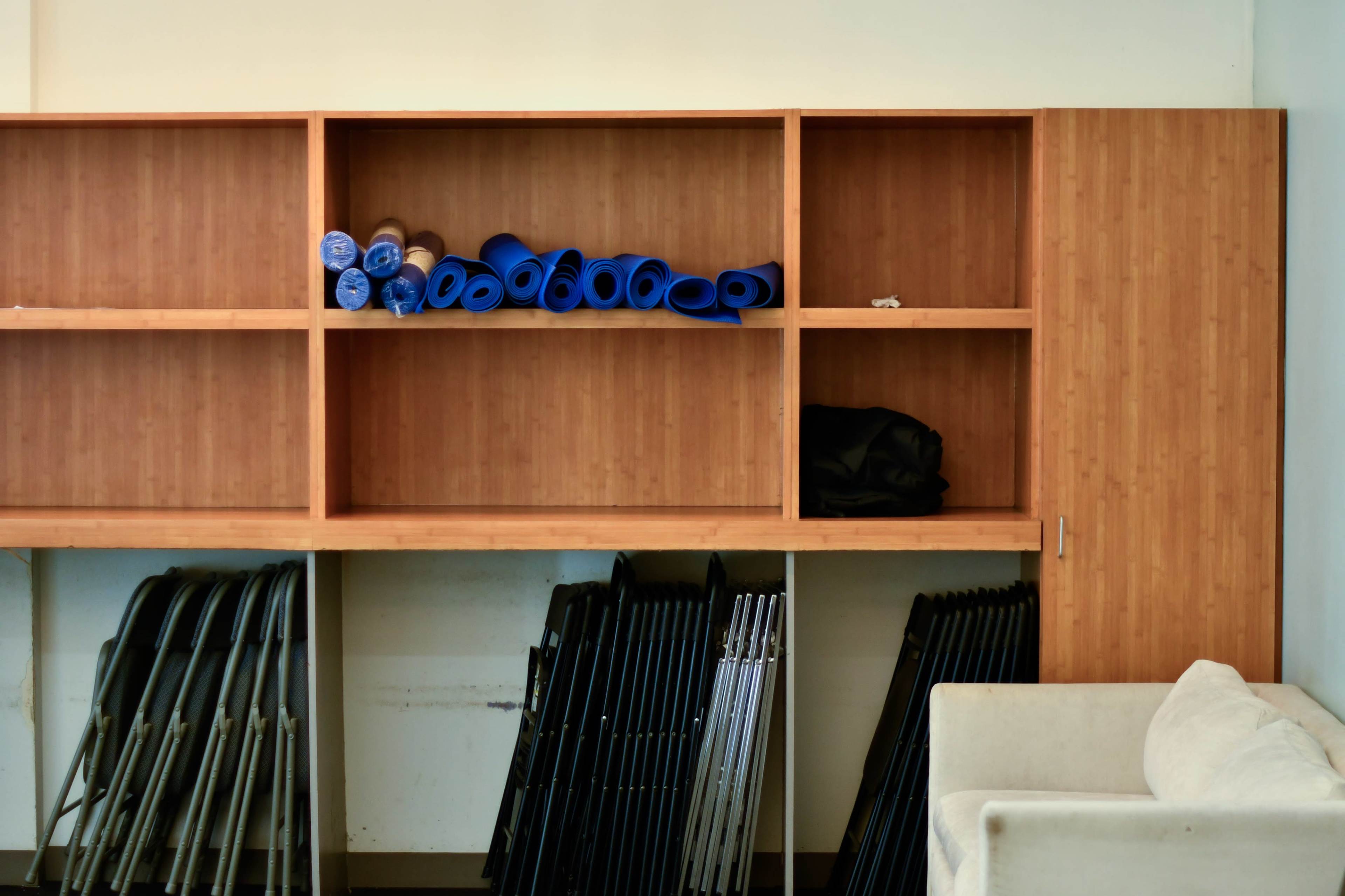 A wooden shelving unit displays rolled blue mats and a black bag, while stacked folding chairs are neatly arranged below.
