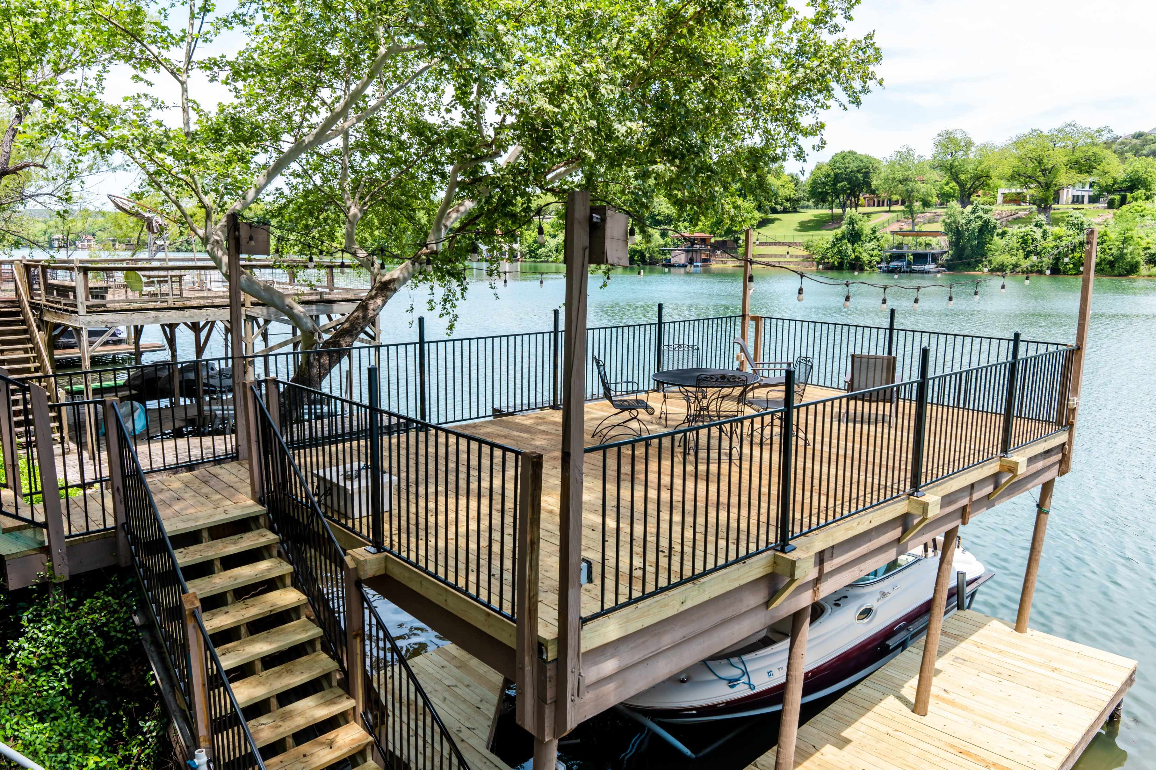 A wooden deck with black railings extends over a lake, featuring a staircase leading down to a dock with a boat.