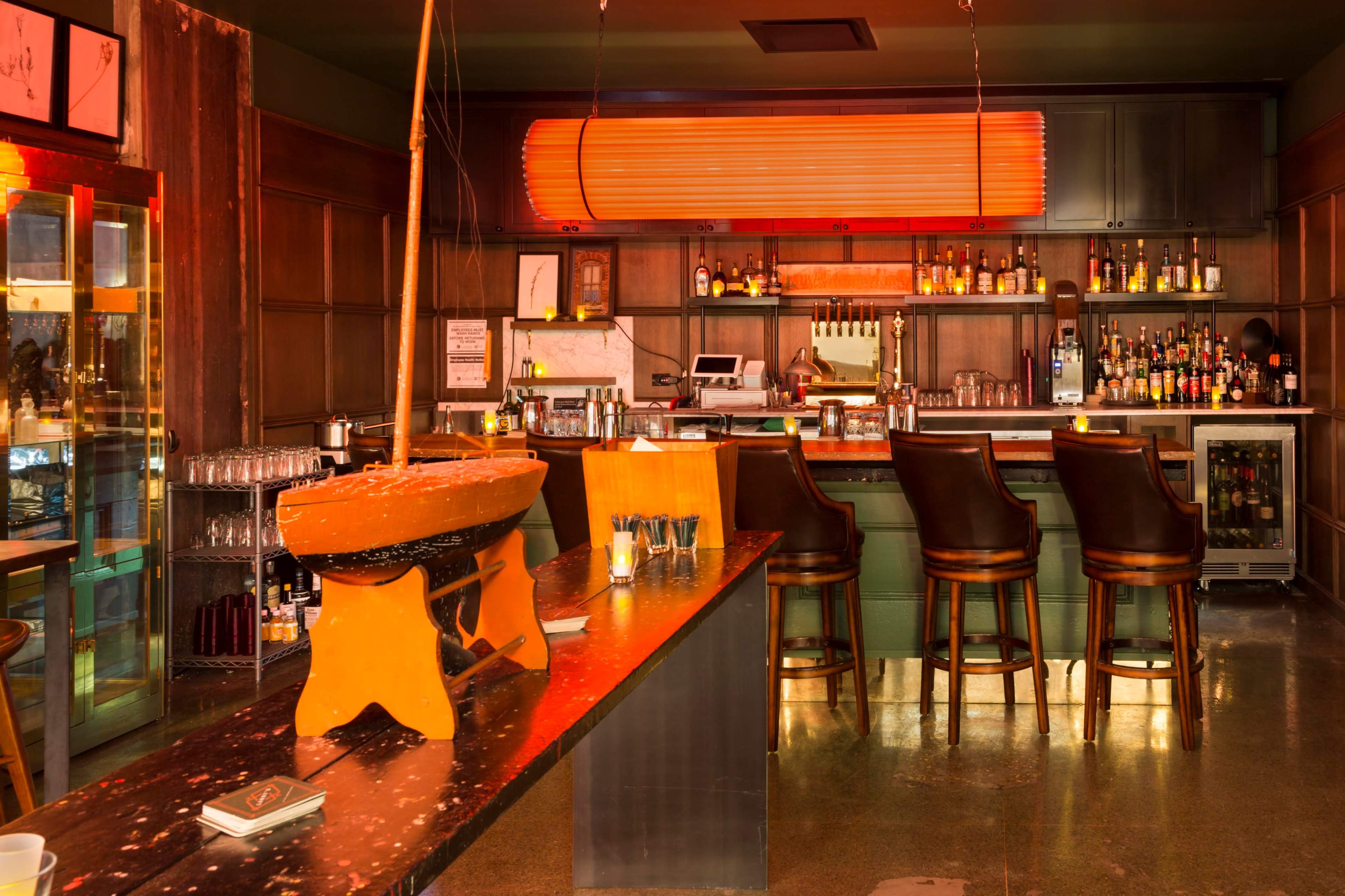 The image shows a stylish bar with wooden furniture, a well-stocked shelf of liquor, and warm orange lighting.