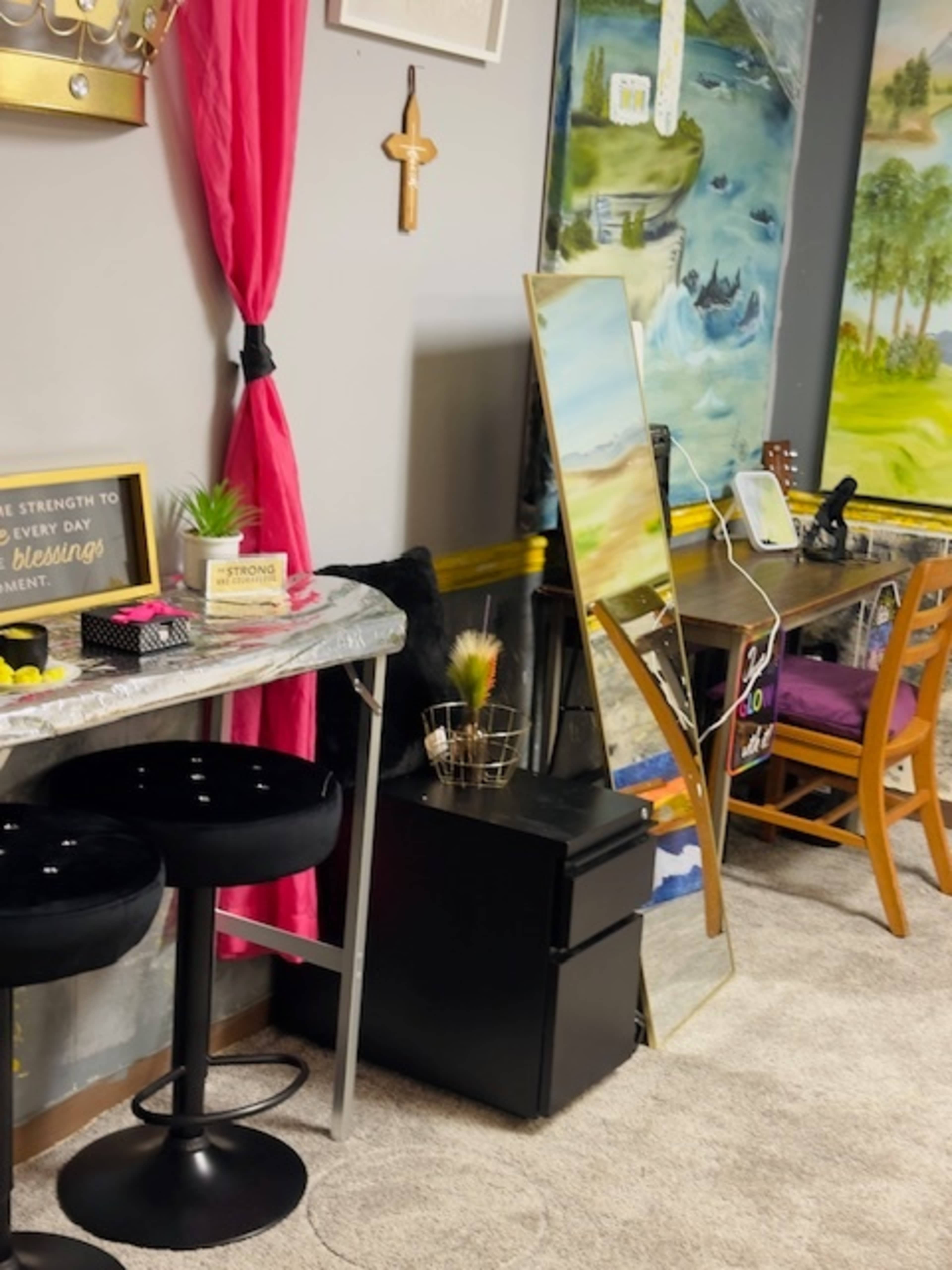 The image shows a room corner with a small table and two black stools, a mirror leaning against the wall, and decorative items including a plant and framed quotes.