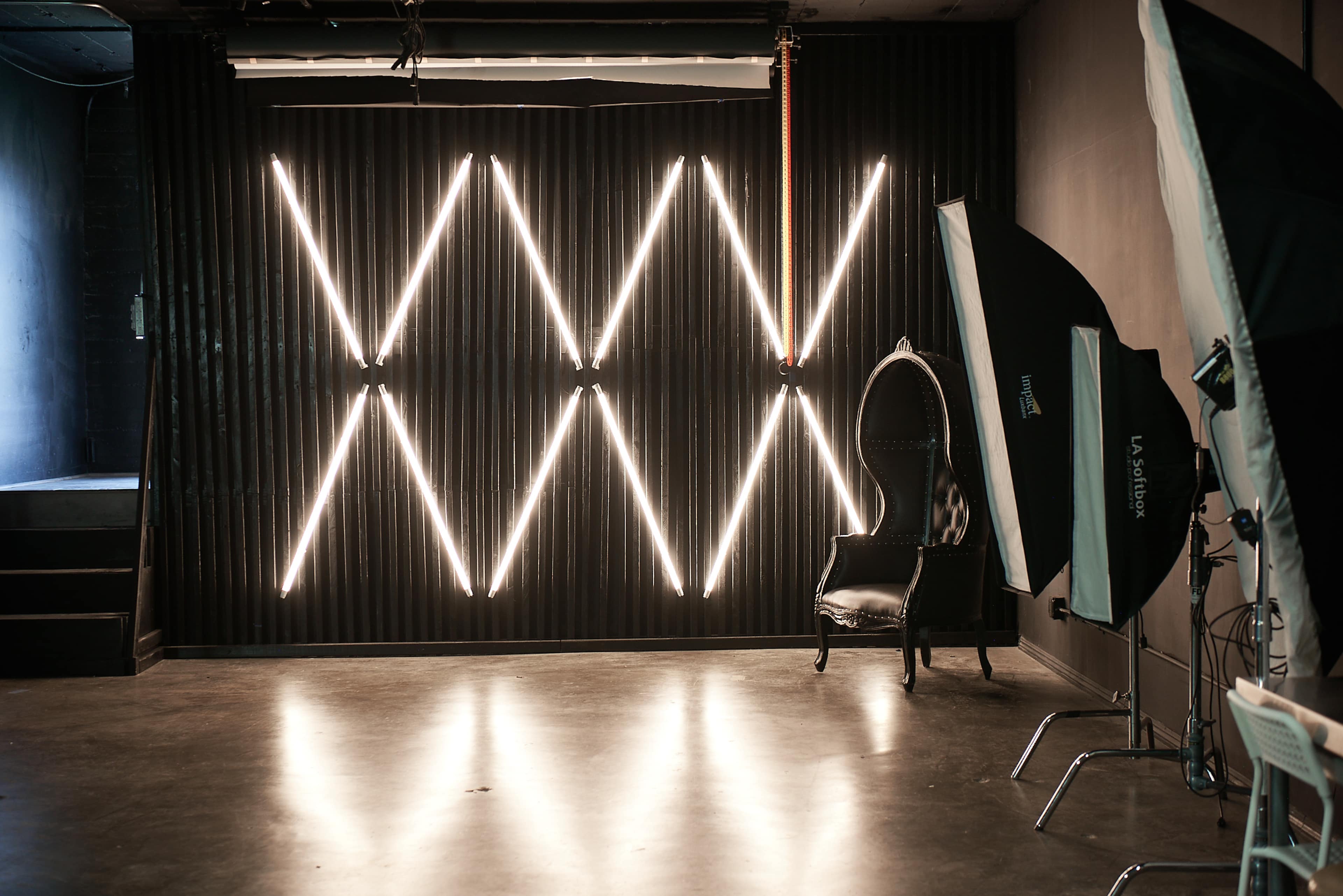 The image shows a photography studio with a dark wall featuring illuminated diagonal neon lines and a vintage-style chair positioned nearby.