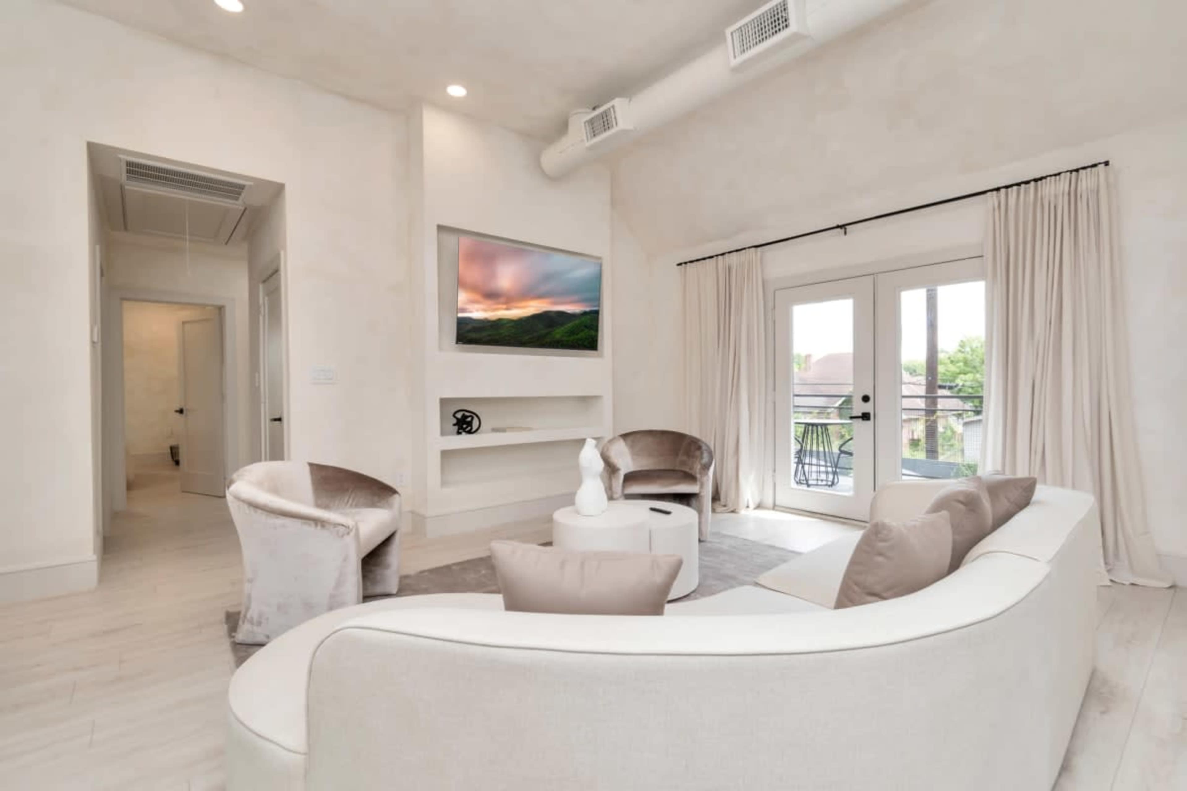 A modern living room with a curved white sofa, two accent chairs, a round coffee table, and a wall-mounted television above a minimalist decorative feature.