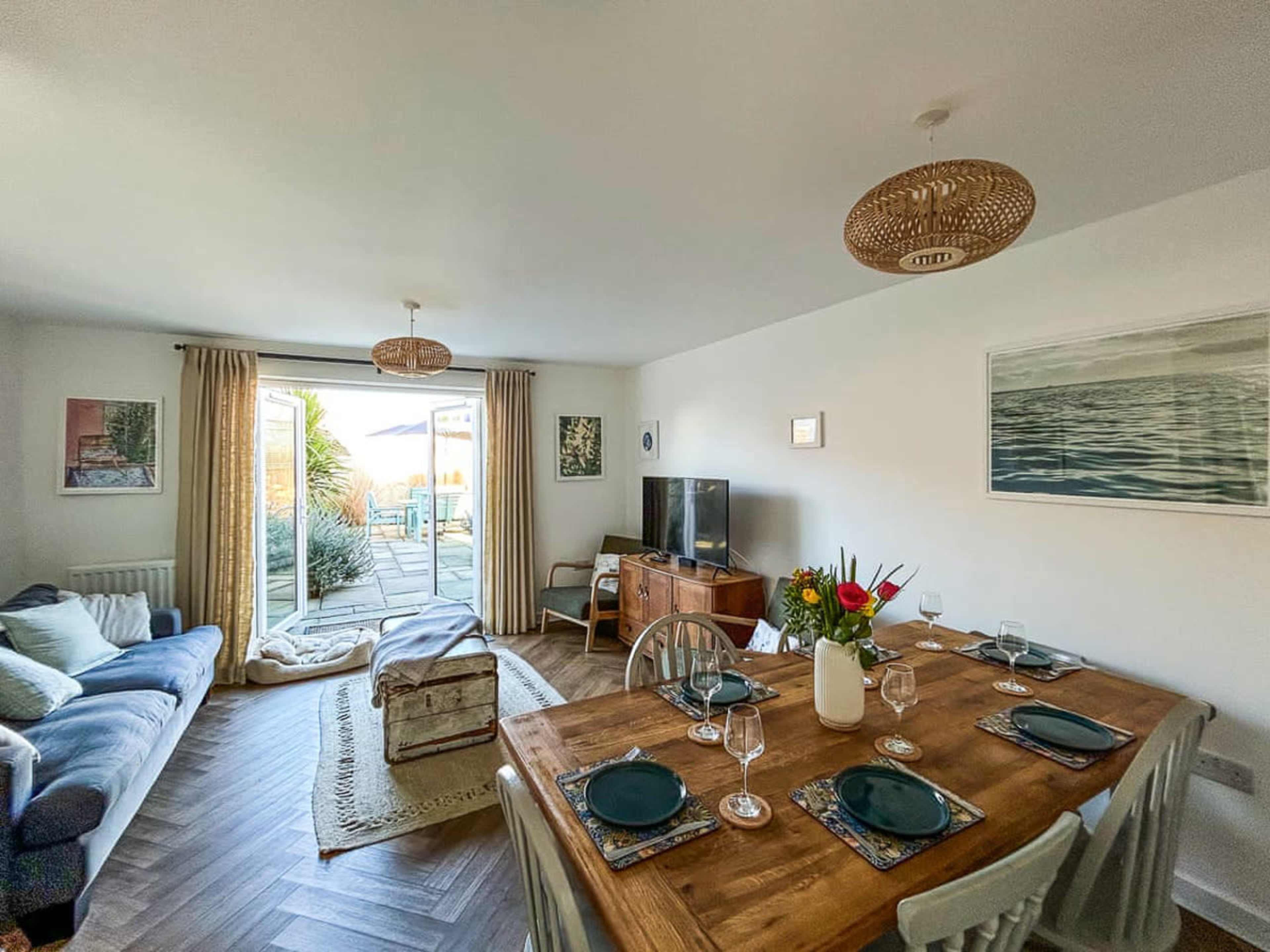 The image shows a bright living and dining area with a wooden table set for four, a blue sofa in the corner, and a view of an outdoor patio through glass doors.