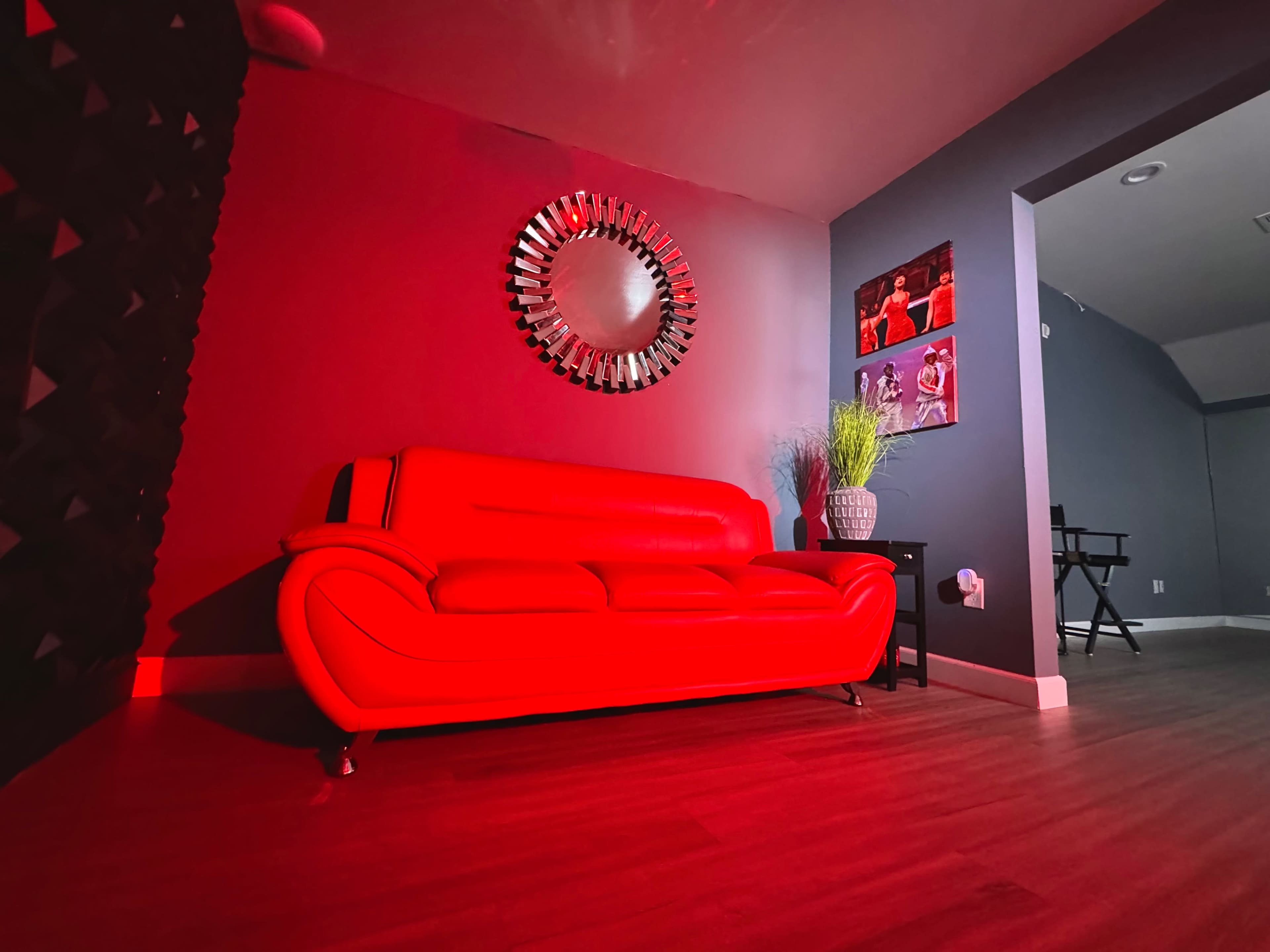 A red sofa is positioned against a wall with a mirror and a plant nearby, in a dimly lit room featuring dark gray walls and a doorway to another area.