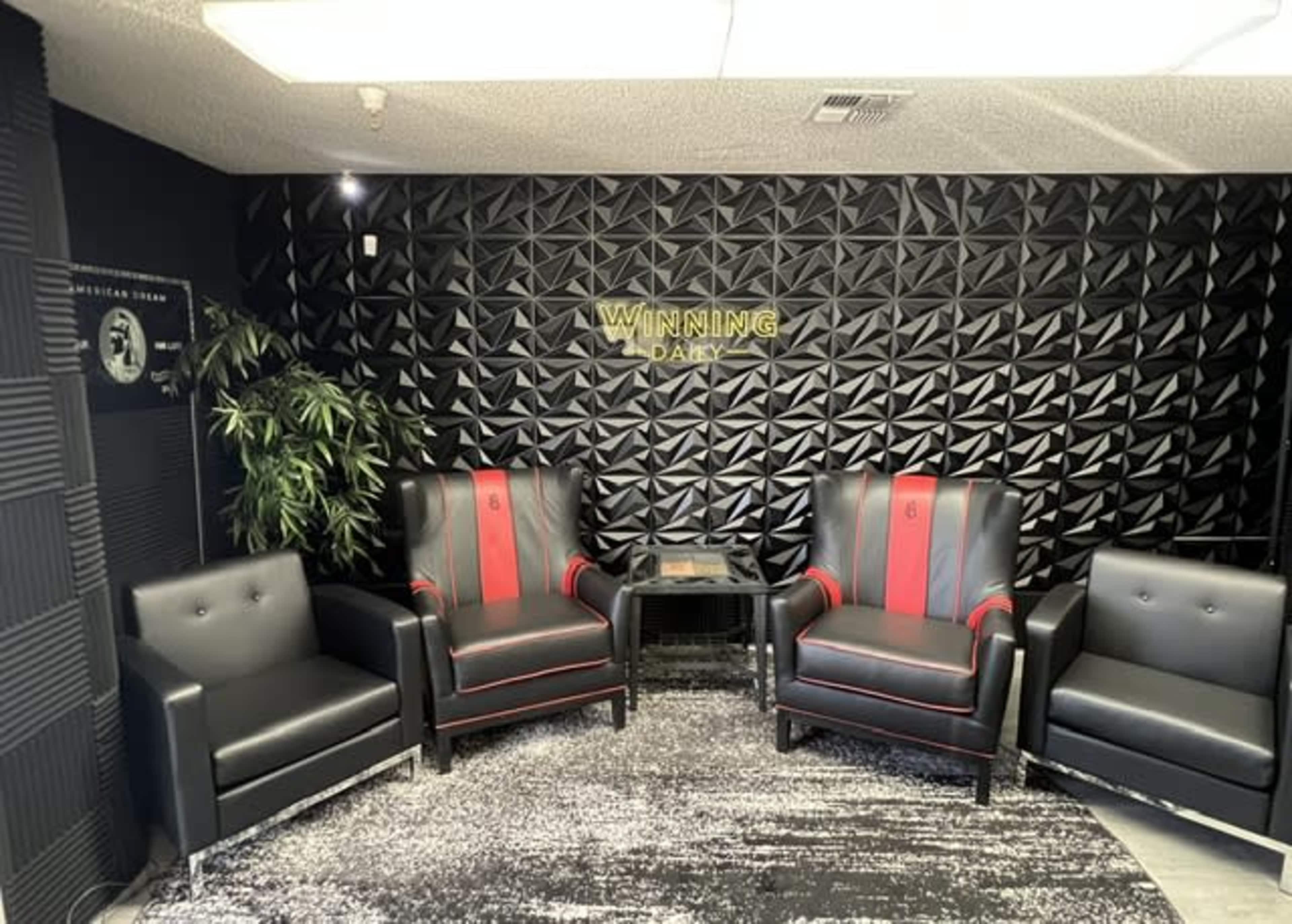 The image shows a modern lounge area featuring four black chairs, two of which have red accents, in front of a textured black wall with the words "WINNING DAILY" in gold lettering.