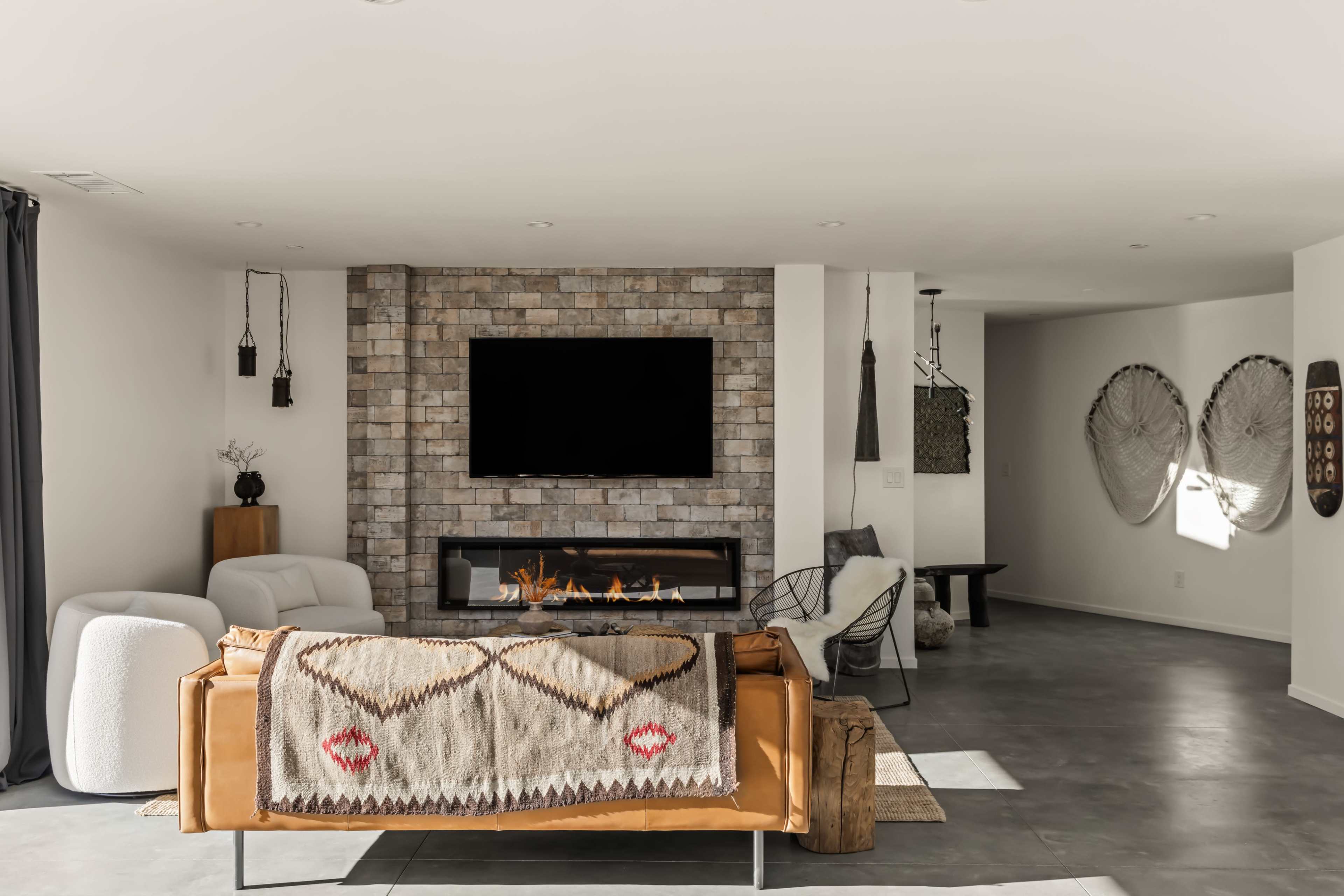 The image shows a modern living room featuring a leather sofa with a blanket, a wall-mounted television, a stone fireplace, and minimalist decor elements.