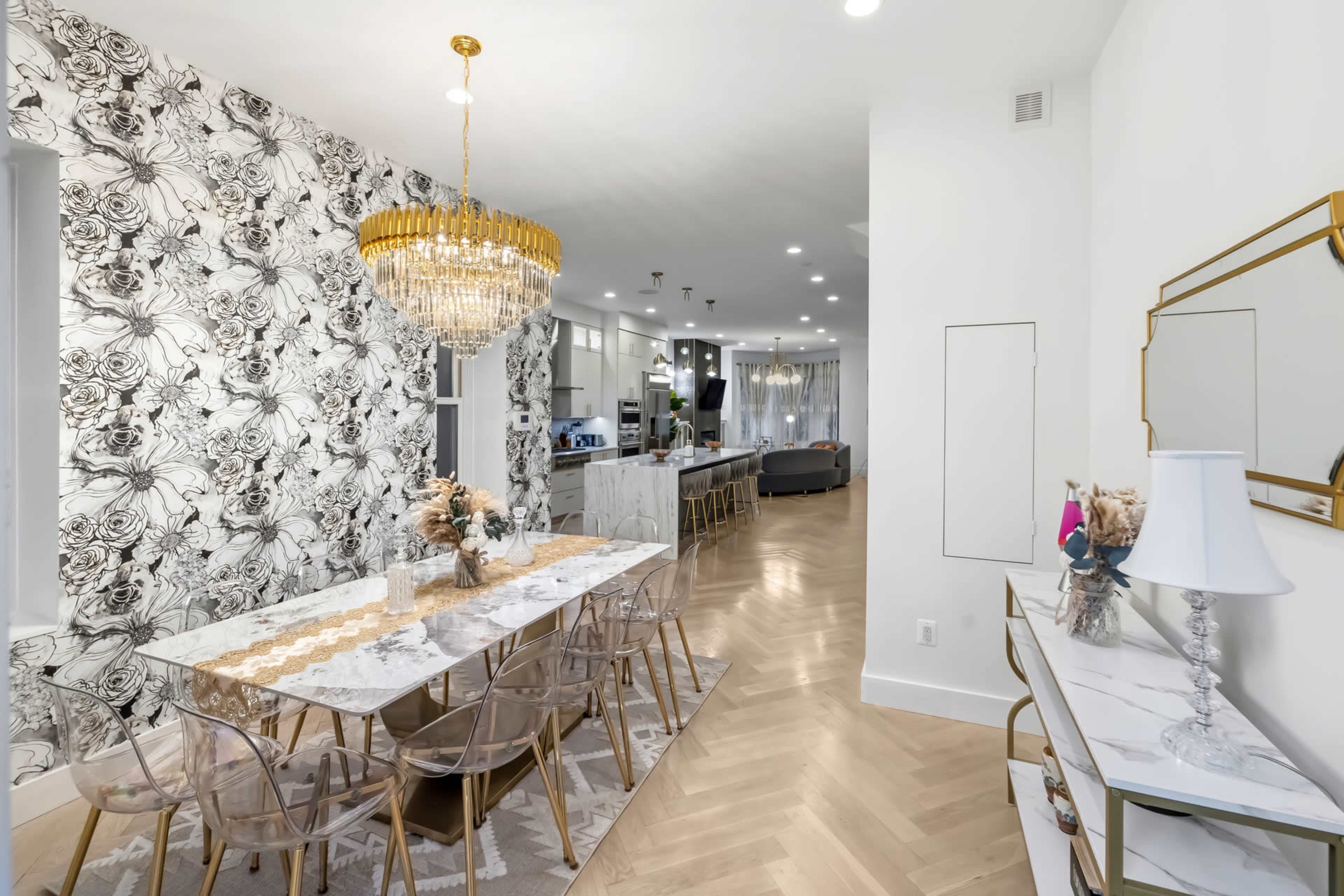 The image shows a modern dining area with a large marble table, transparent chairs, a decorative chandelier, and a floral wallpaper backdrop, leading into an open kitchen and living space.