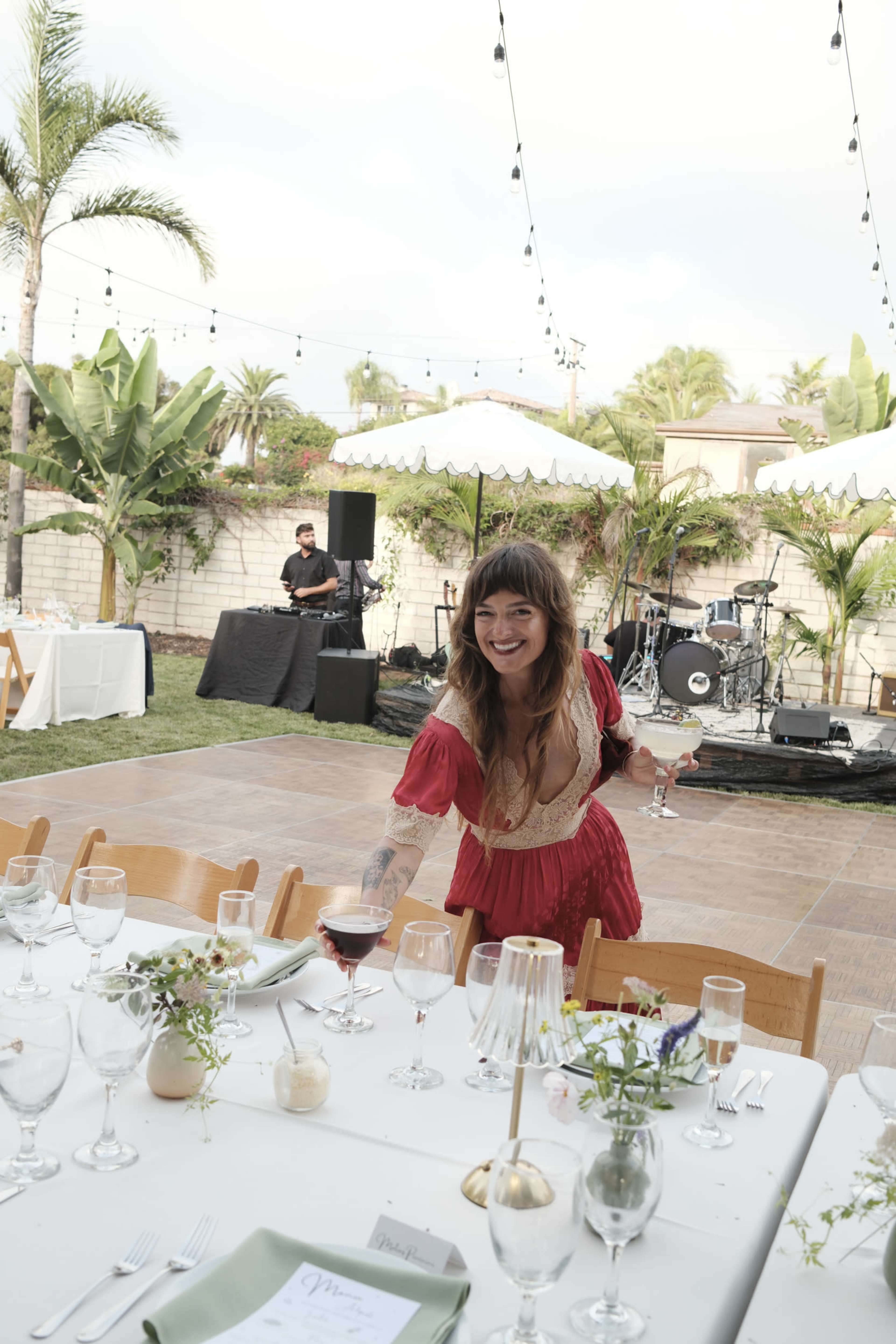 A woman in a red dress smiles while setting a table at an outdoor event space with palm trees and a stage in the background.