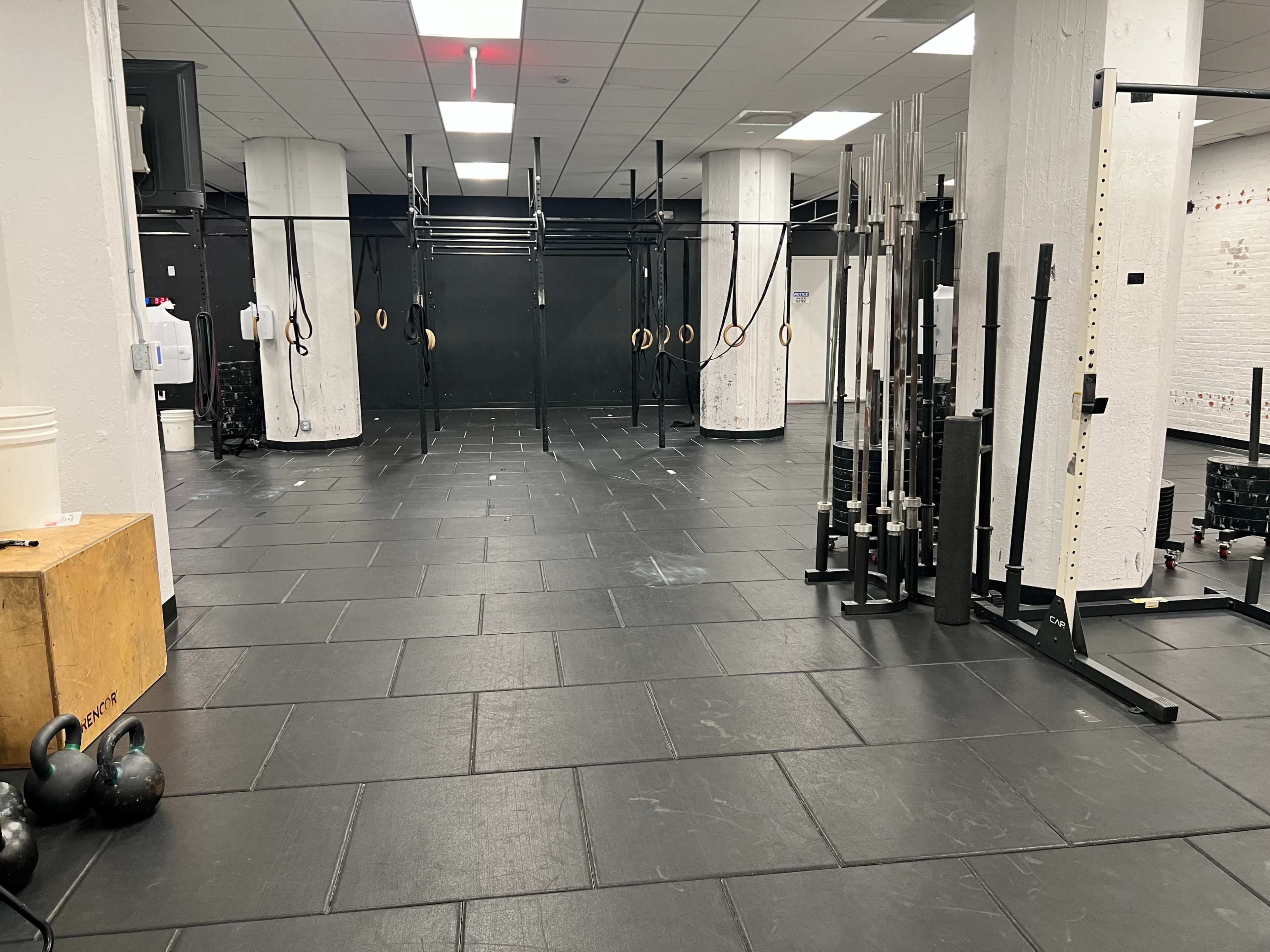 A spacious gym area with various exercise equipment, including kettlebells, a wooden box, and weightlifting bars, arranged on black rubber flooring.