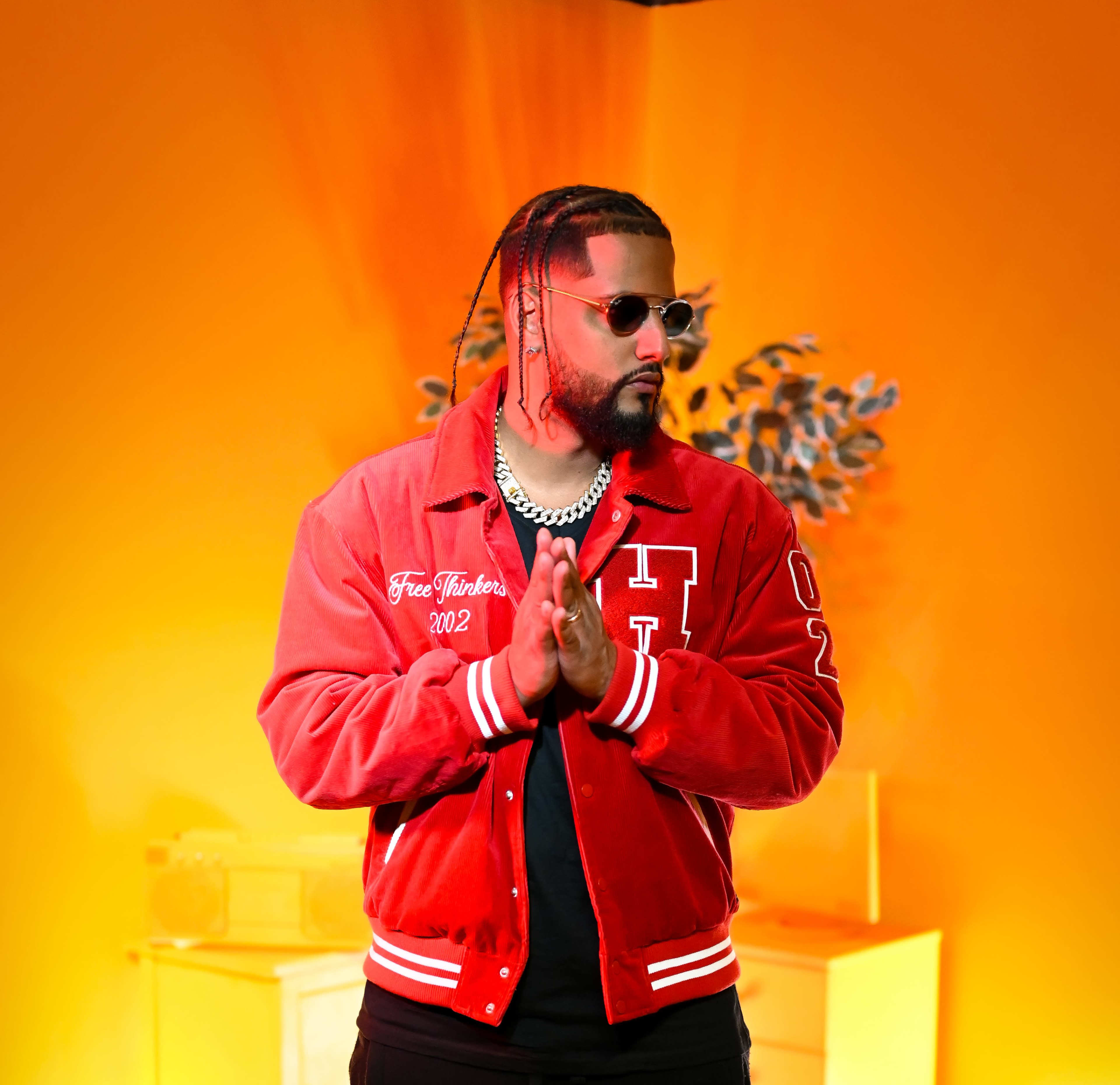 A man wearing a red jacket and sunglasses stands with his hands clasped in front of a bright orange background.