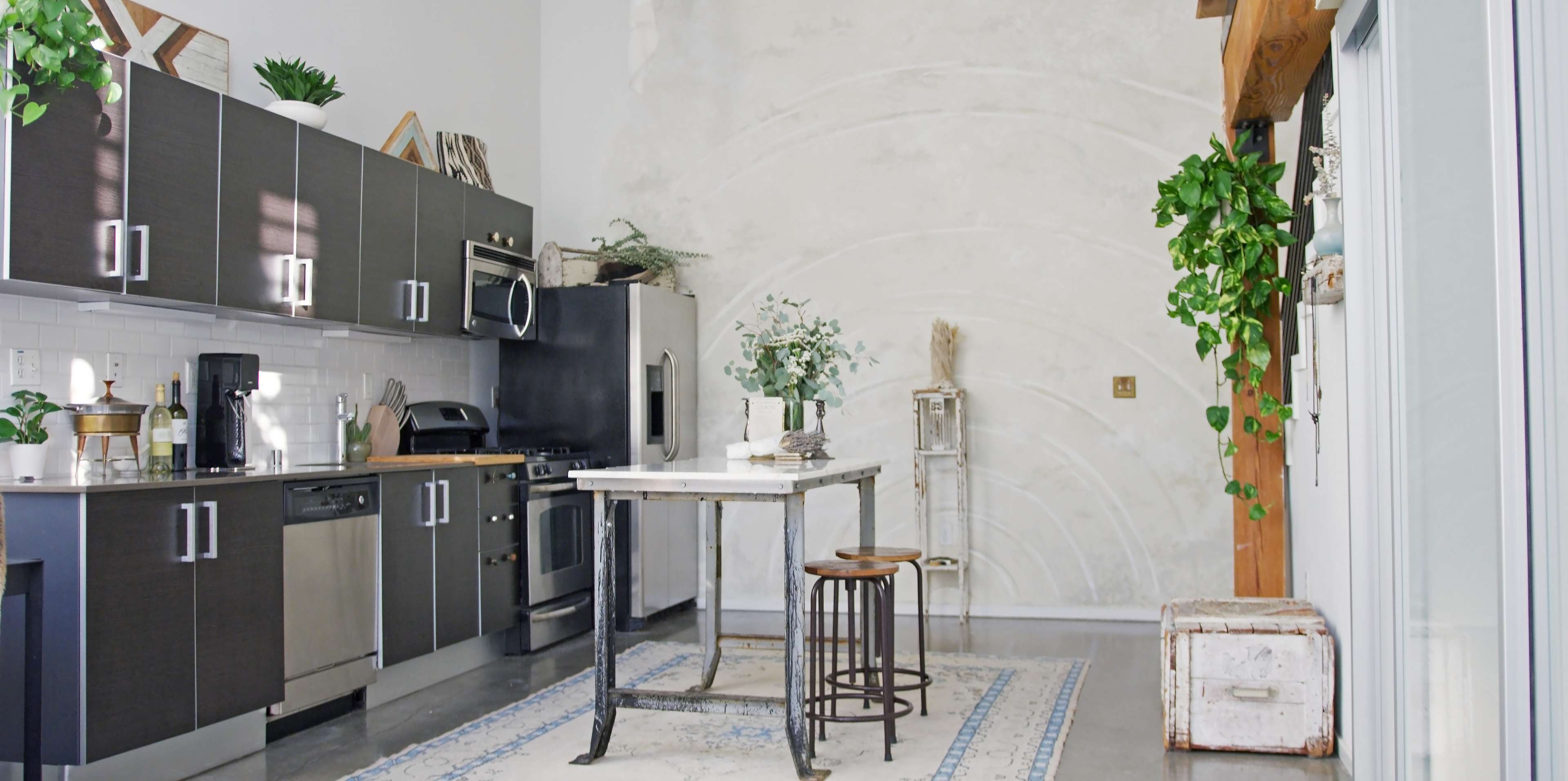 A modern kitchen with dark cabinetry, stainless steel appliances, a small dining table, and a potted plant in the corner.