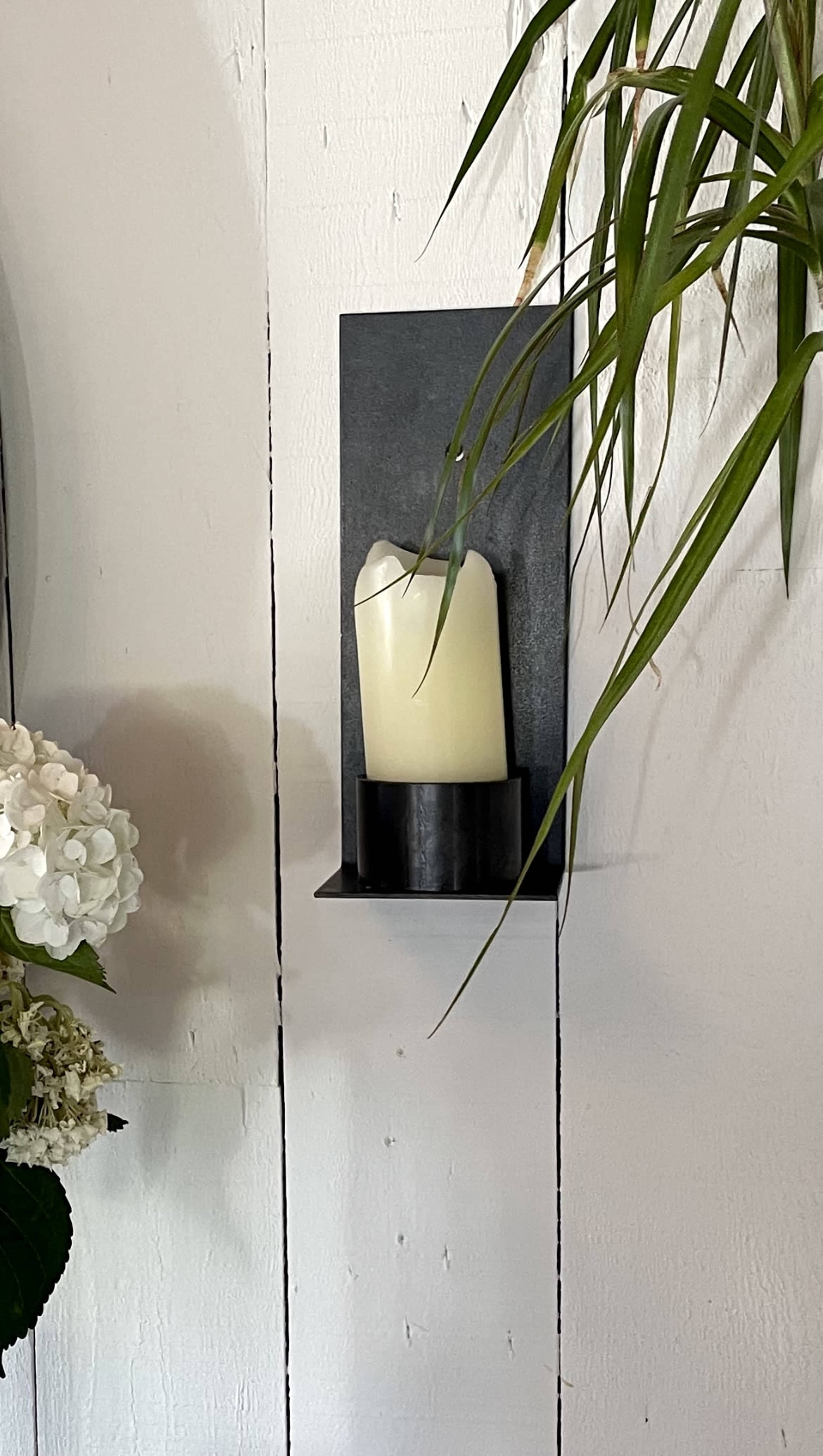 A white candle is positioned on a black metal wall sconce against a backdrop of white wooden planks, with a green plant hanging nearby.